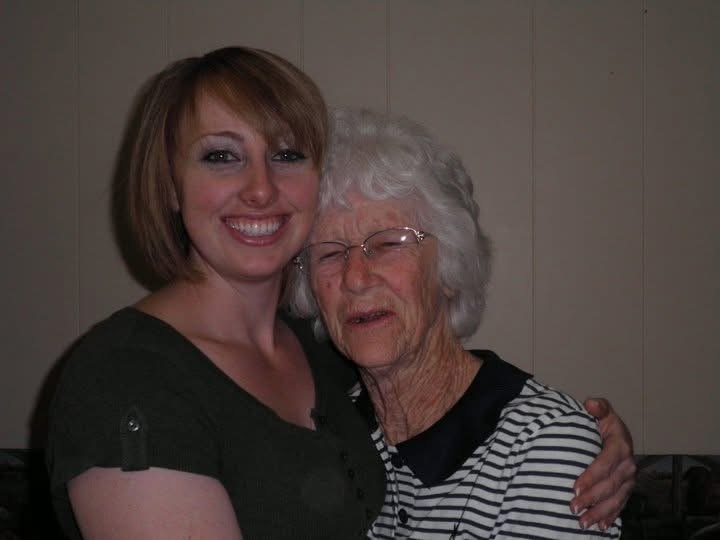 Two women hugging and smiling, an older woman with gray hair and glasses wearing a black and white striped shirt, and a younger woman with brown hair wearing a dark shirt.