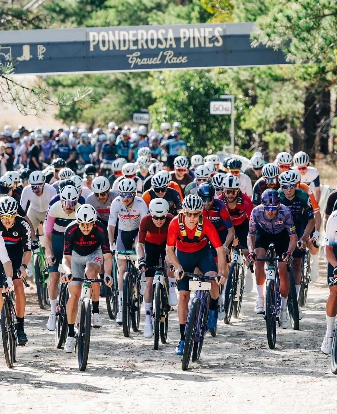 Cyclists start a race at the Ponderosa Pines trail, wearing helmets and colorful athletic gear.