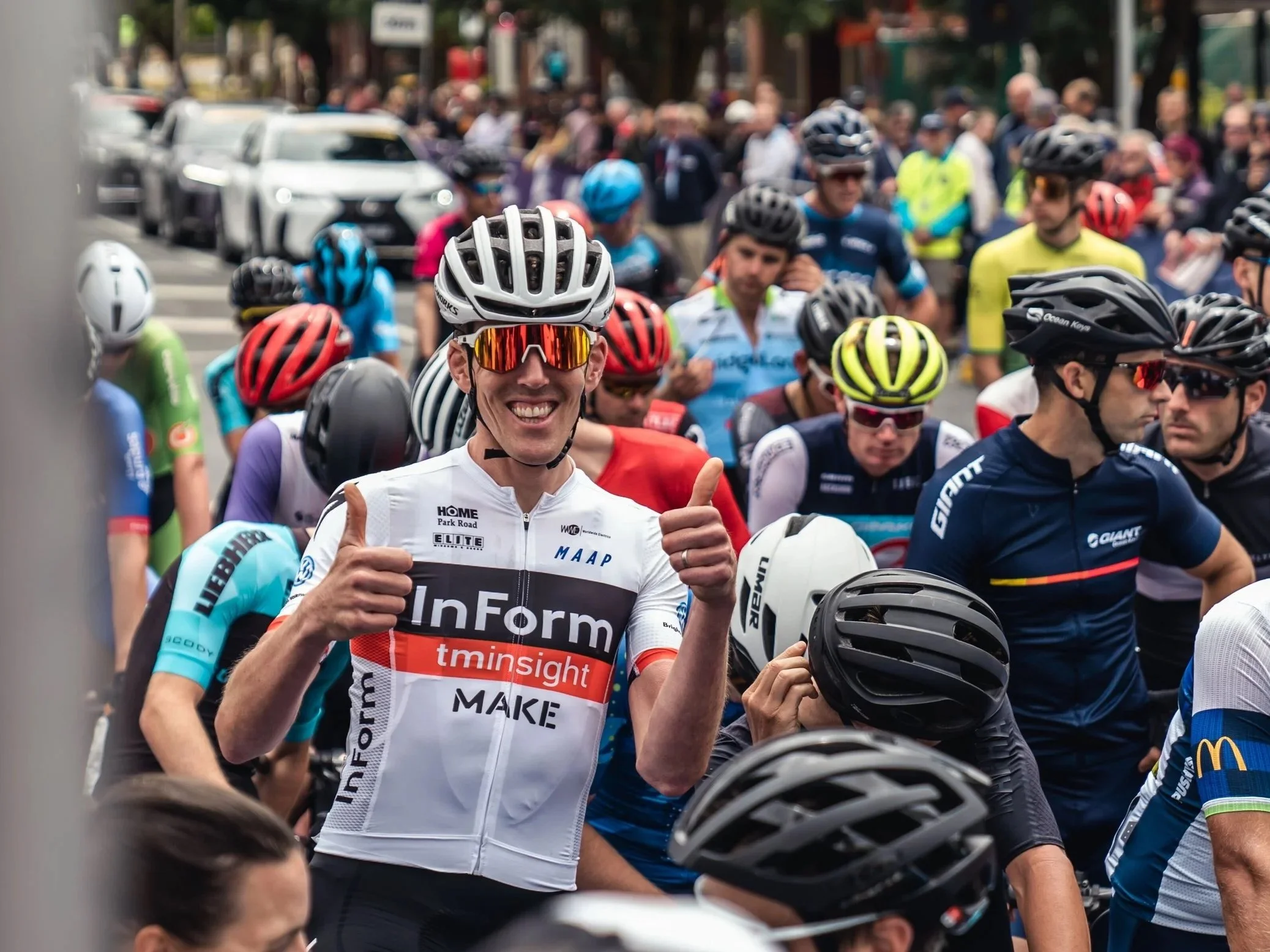 A group of cyclists in colorful jerseys and helmets gathers for a race, with a smiling cyclist in white giving a thumbs-up in the foreground.