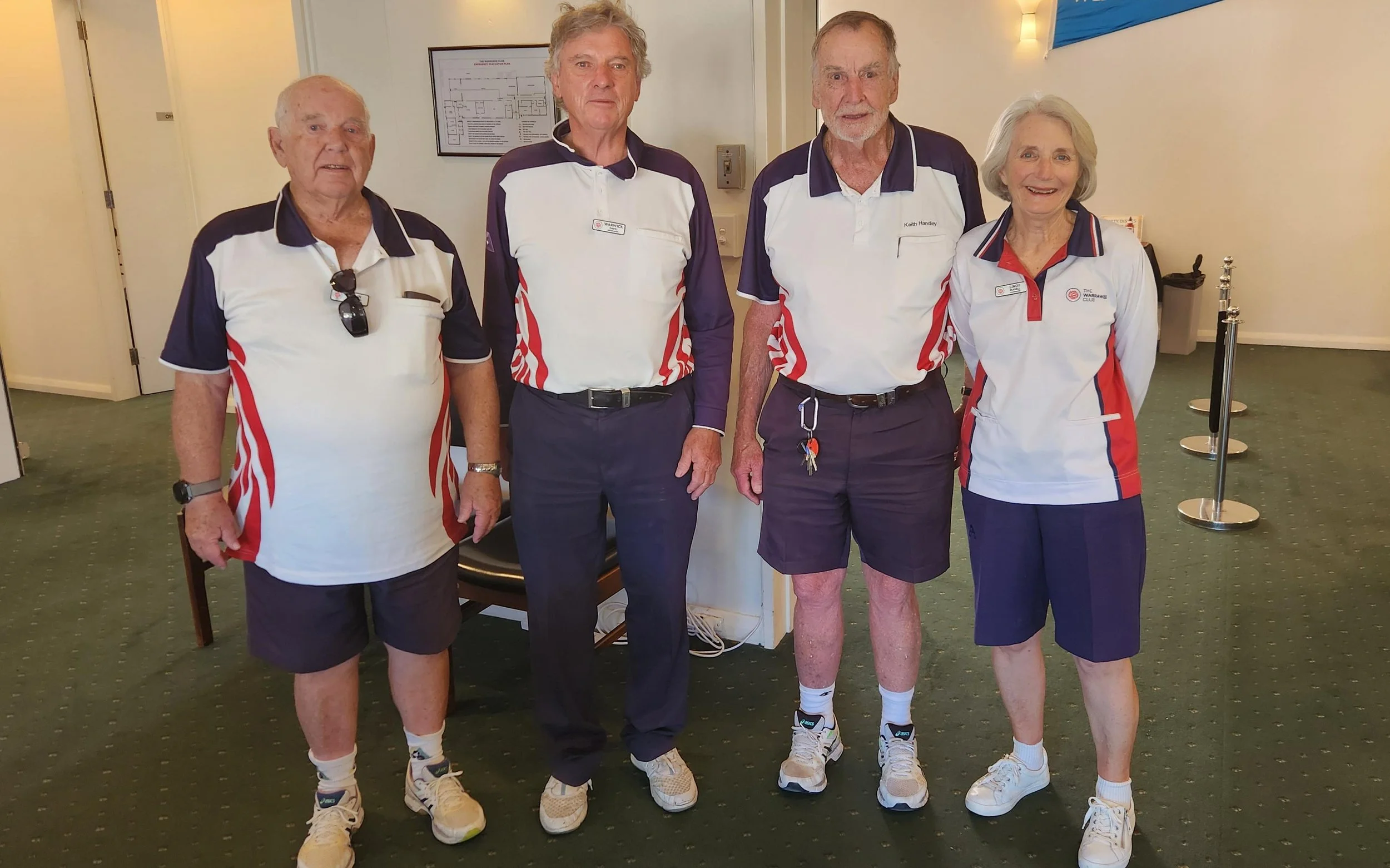 Four older adults in white and navy sports uniforms standing indoors on a green carpet, smiling for a group photo.