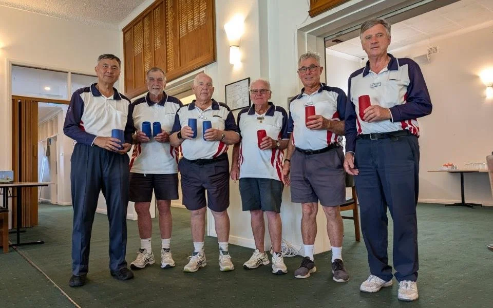 Six older men wearing matching white, navy, and red sports shirts, shorts or pants, holding cups or mugs, standing indoors in front of a wall with a framed certificate or diploma.