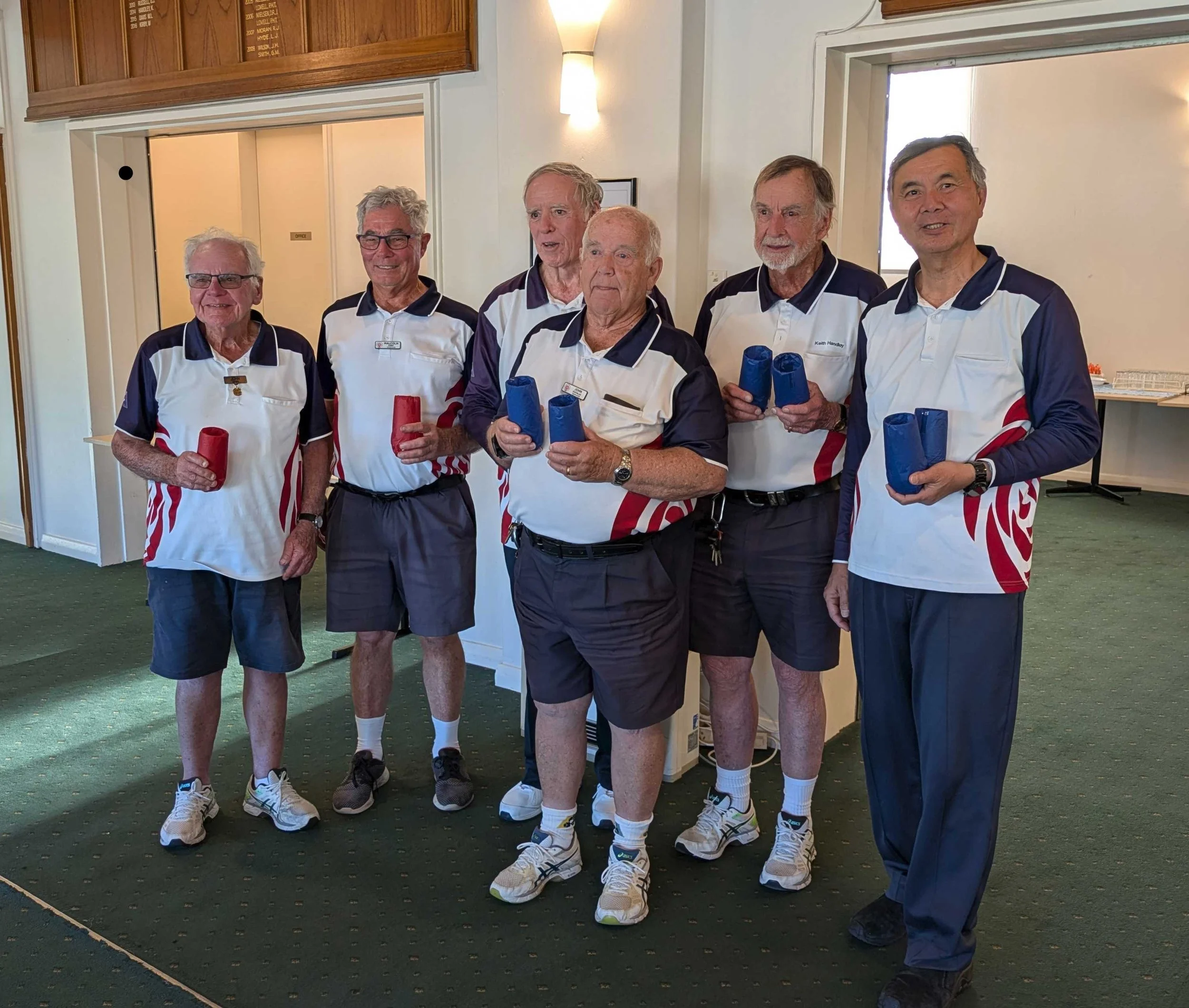 A group of six elderly men wearing matching sports uniforms standing indoors, holding cylindrical objects, likely trophies or awards, smiling for the photo.