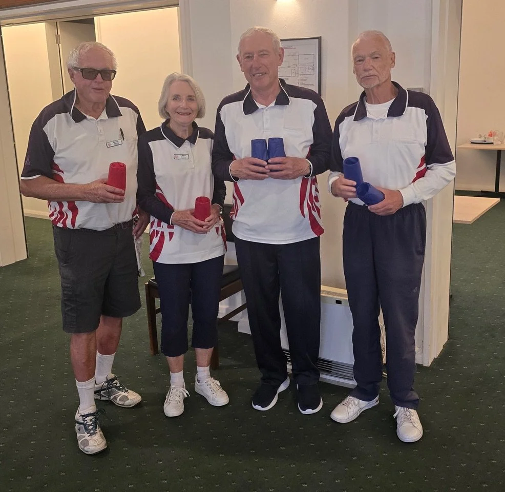 four elderly individuals standing indoors in sports jerseys, holding stacked cups, with a green carpeted floor and a wall-mounted framed picture in the background.