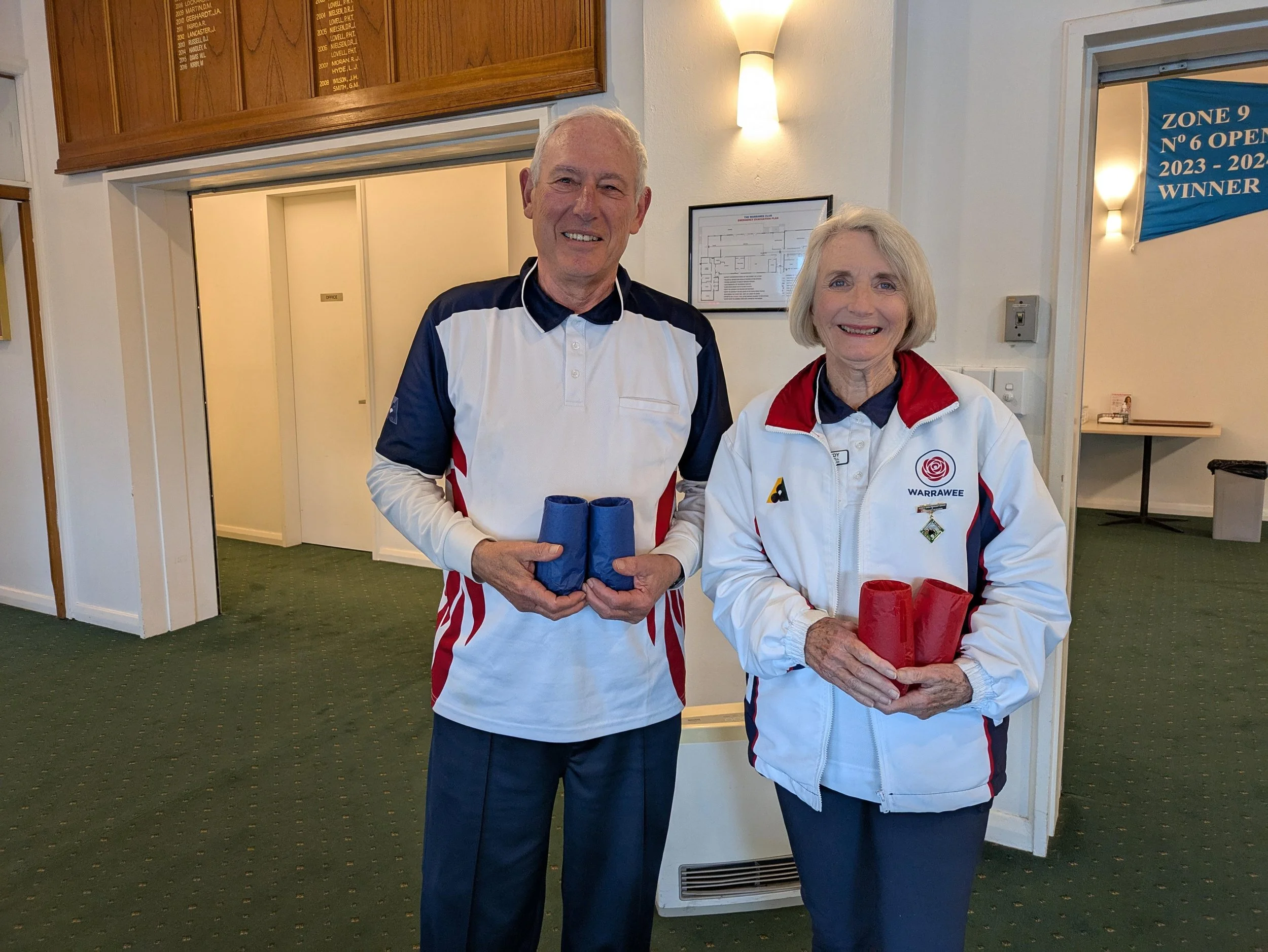 A man and a woman are smiling and holding red and blue bowling pins, standing in an indoor sports facility with green carpet and white walls.