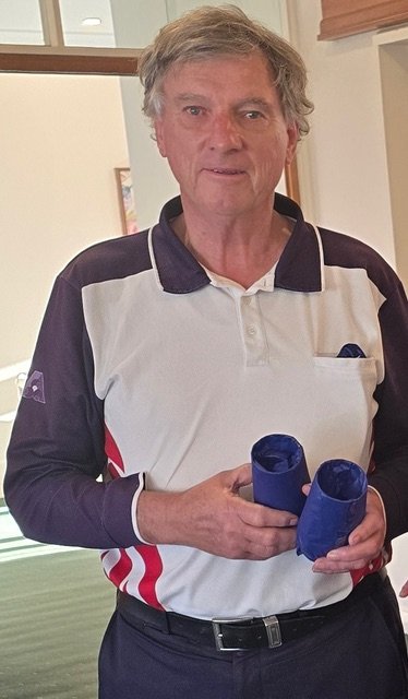 Older man with gray hair wearing a white and navy polo shirt, holding blue rolled-up items, standing indoors.