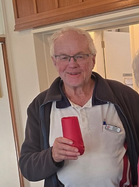 An elderly man smiling and holding a red cup in his right hand. He is wearing glasses, a white polo shirt, and a dark jacket. The background shows a wooden cabinet and part of a doorway.