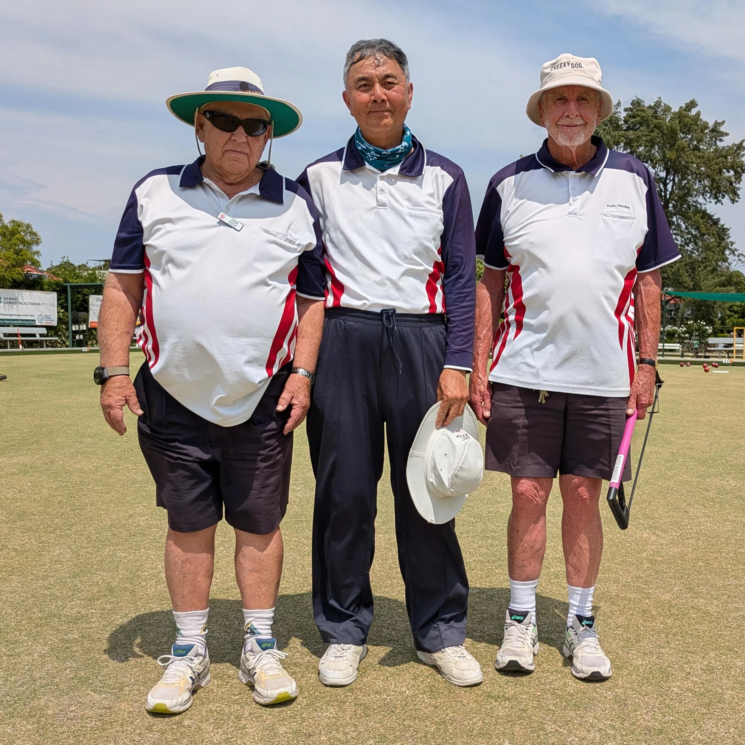 Three senior men standing on a lawn bowls green, wearing matching white and navy sports shirts, with two of them holding hats and one holding a lawn bowls rake. They appear to be participating in lawn bowls.