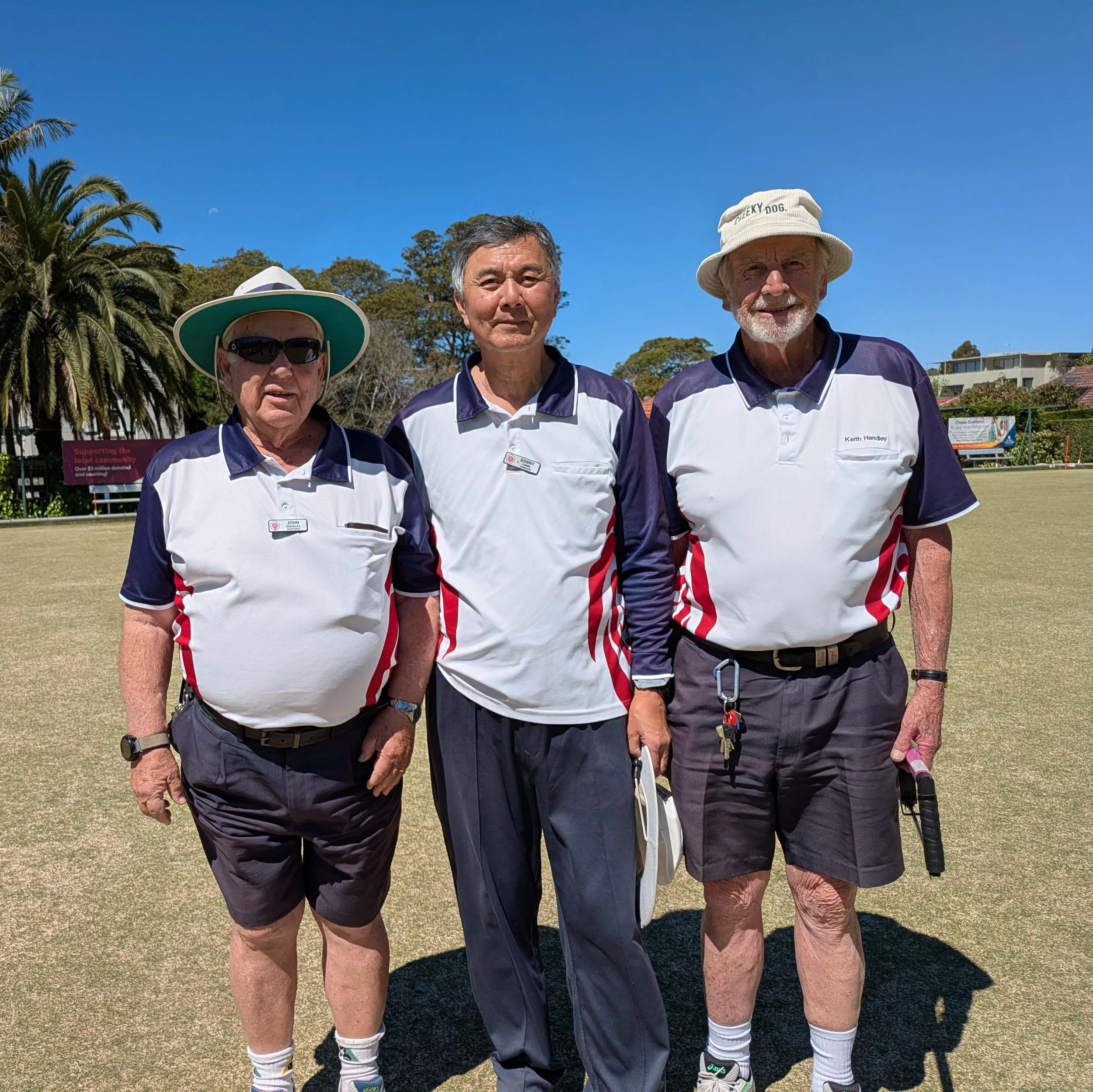 Three elderly men standing on a golf course, wearing matching white and navy blue sports shirts with red accents, and hats, under a clear blue sky with palm trees in the background.