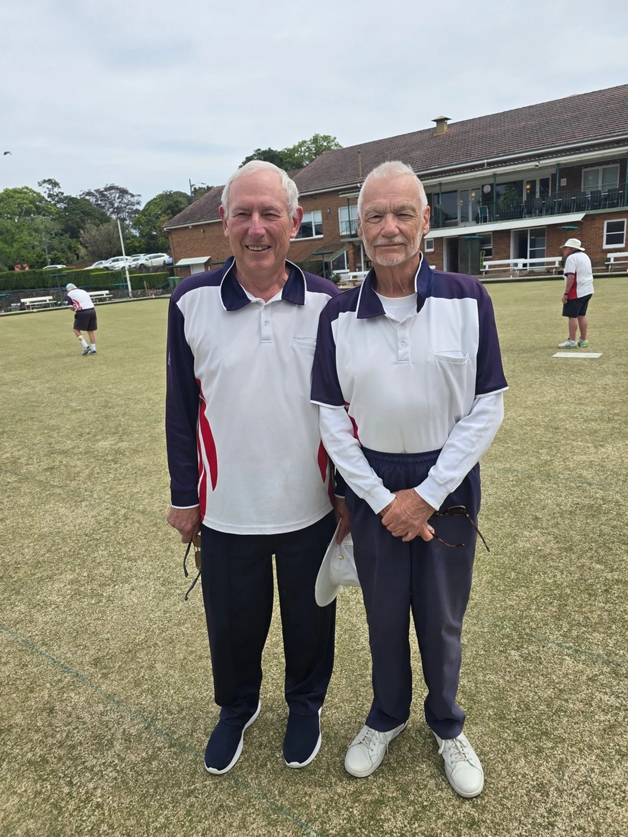 Two elderly men standing on a lawn bowling green, wearing matching white and navy sports shirts, with a building and other players in the background.