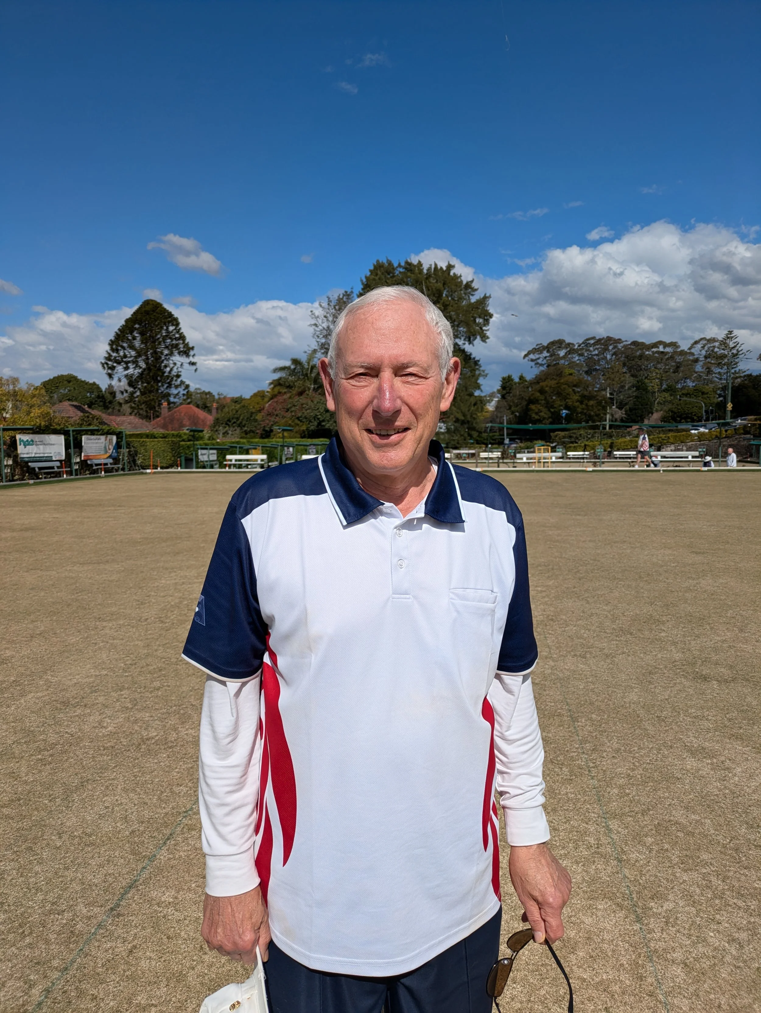 An older man with short gray hair, wearing a blue and white sports shirt with red accents, standing on a croquet court under a partly cloudy sky, holding sunglasses in his right hand.