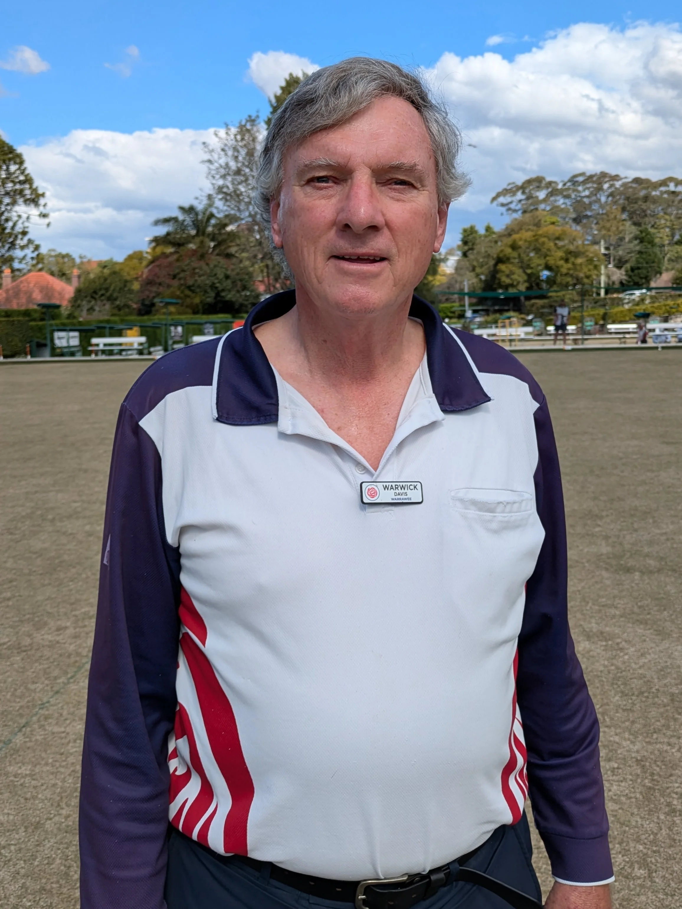 A middle-aged man with gray hair standing on a lawn bowling green, wearing a white and navy blue sports shirt with the name tag "Warwick Davis".
