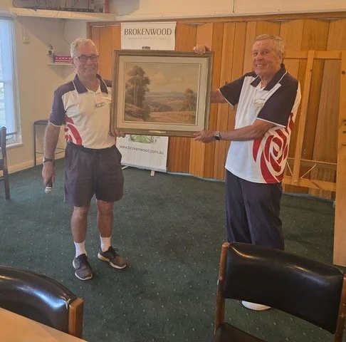 Two men in golf shirts and shorts holding a framed landscape painting in a room with wood paneling and a 'Brokenwood' sign in the background.