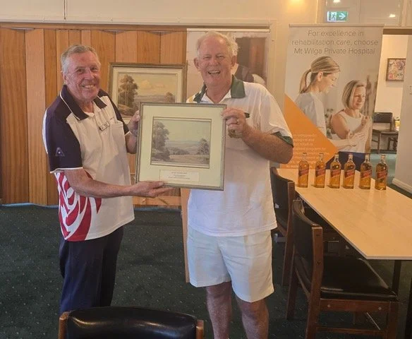 Two smiling men, one holding a framed landscape painting, in a wood-paneled room with a booth in the background displaying bottles and a poster.