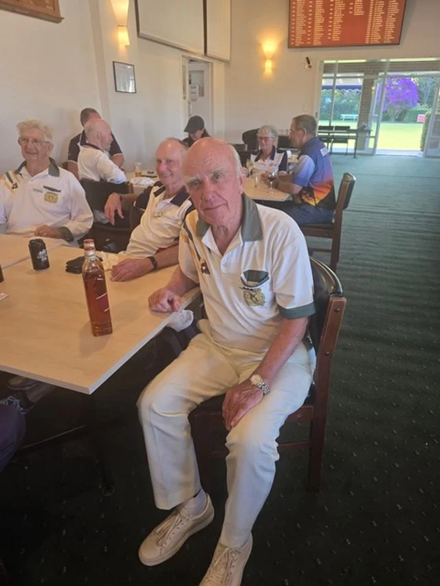 A group of older men in golf uniforms sitting in a golf club or banquet room, some smiling, with drinks on the table. Through the window, a golf course and purple flowering bush are visible.