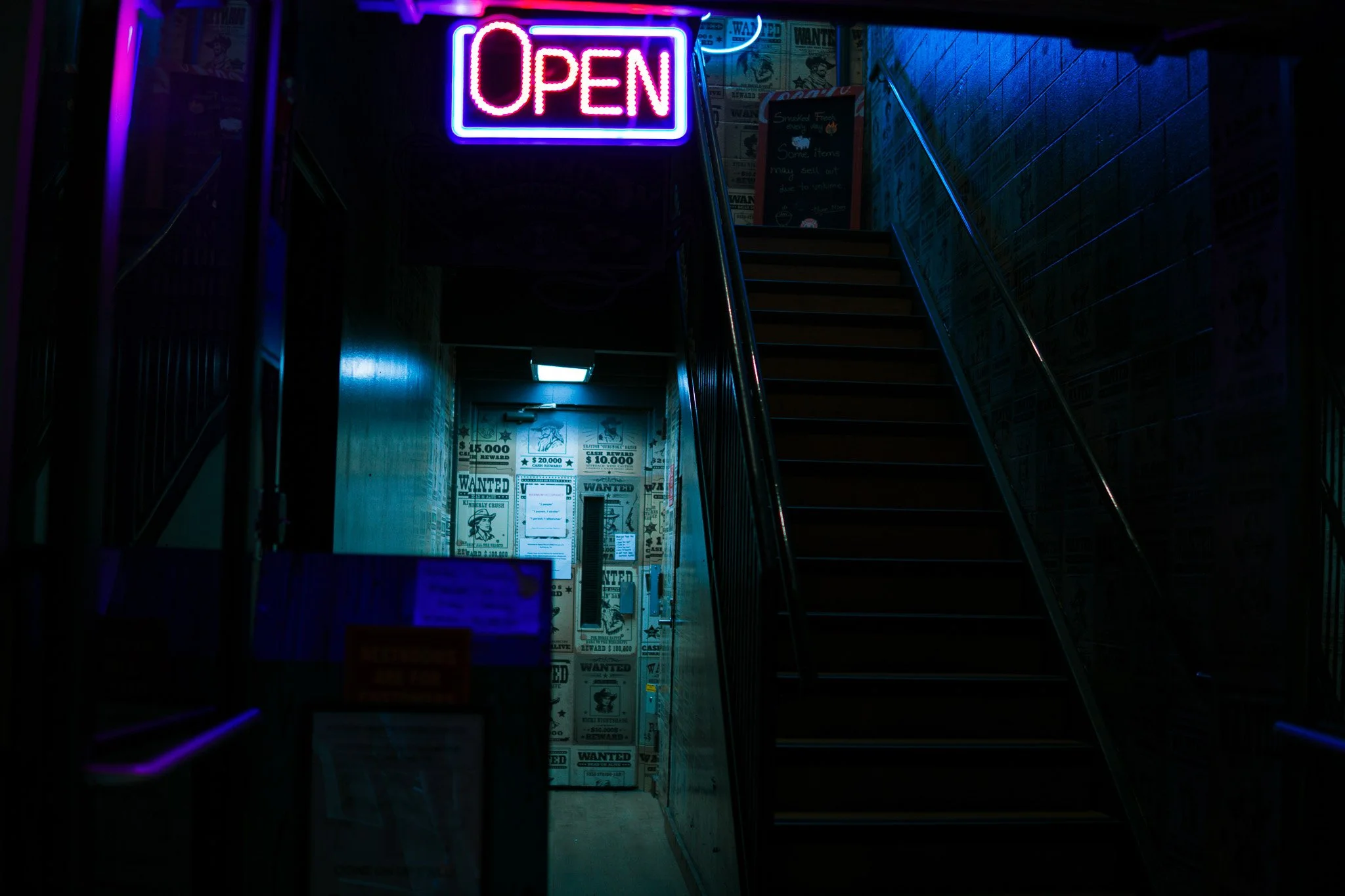 Neon sign with the word 'OPEN' in pink and blue lights at the top of a staircase inside a dimly lit establishment.
