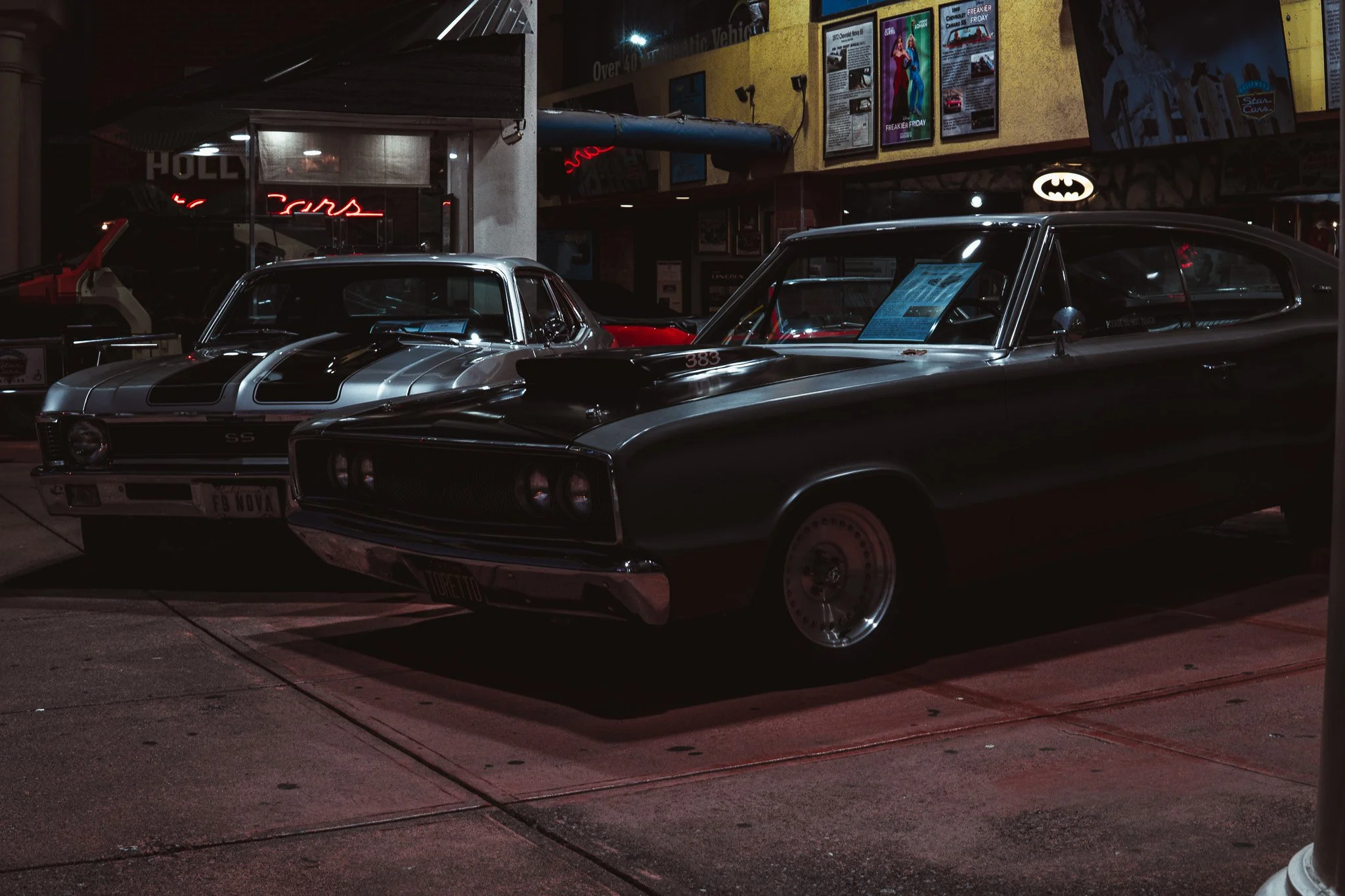 Nighttime scene of two vintage cars parked outside a building with bright signs and posters, including a Batman logo and movie posters, illuminated in the dark.