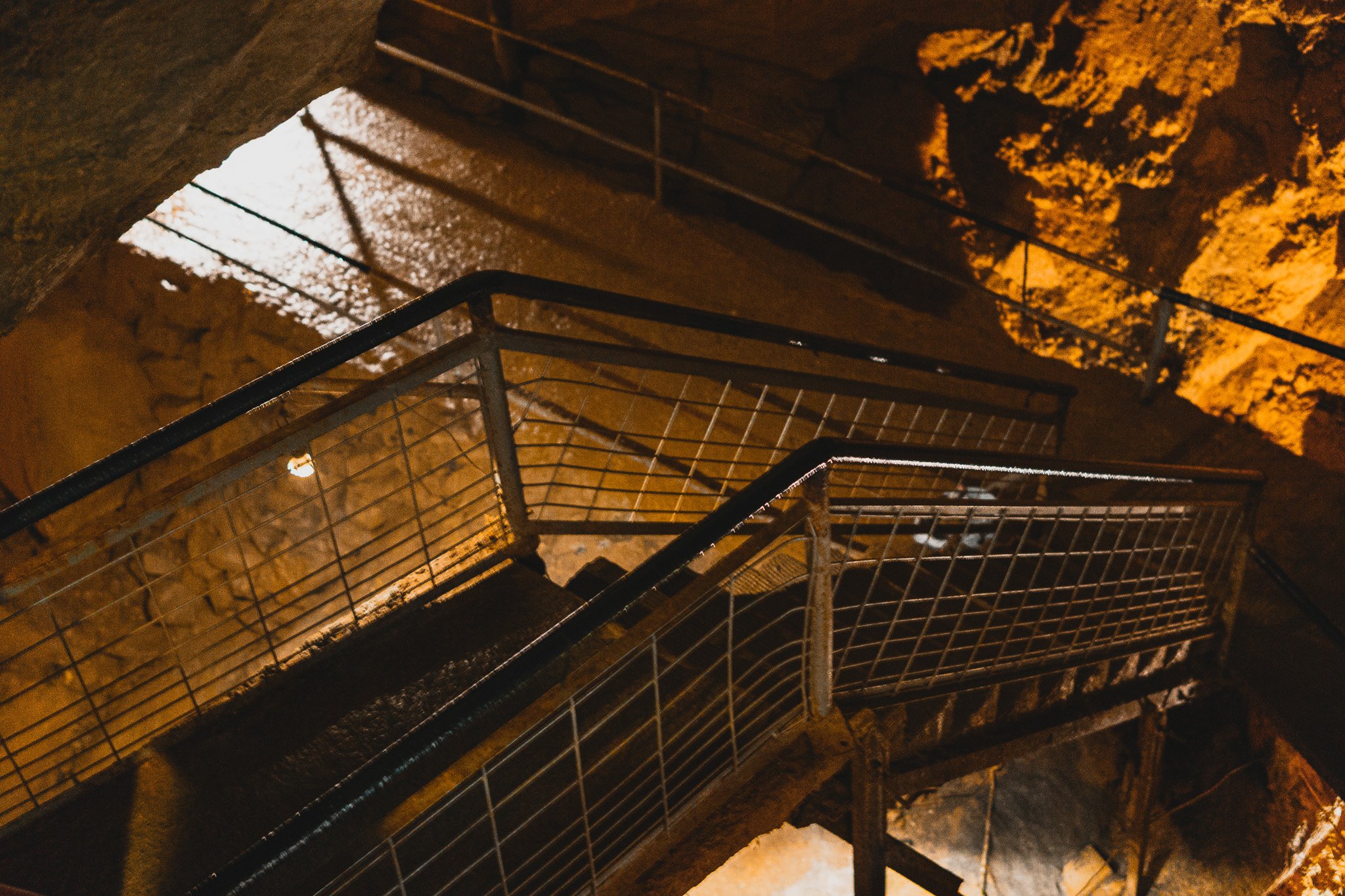 View from the top of a staircase looking down, with metal railings and stone steps illuminated by nearby lights, and a rocky wall in the background.