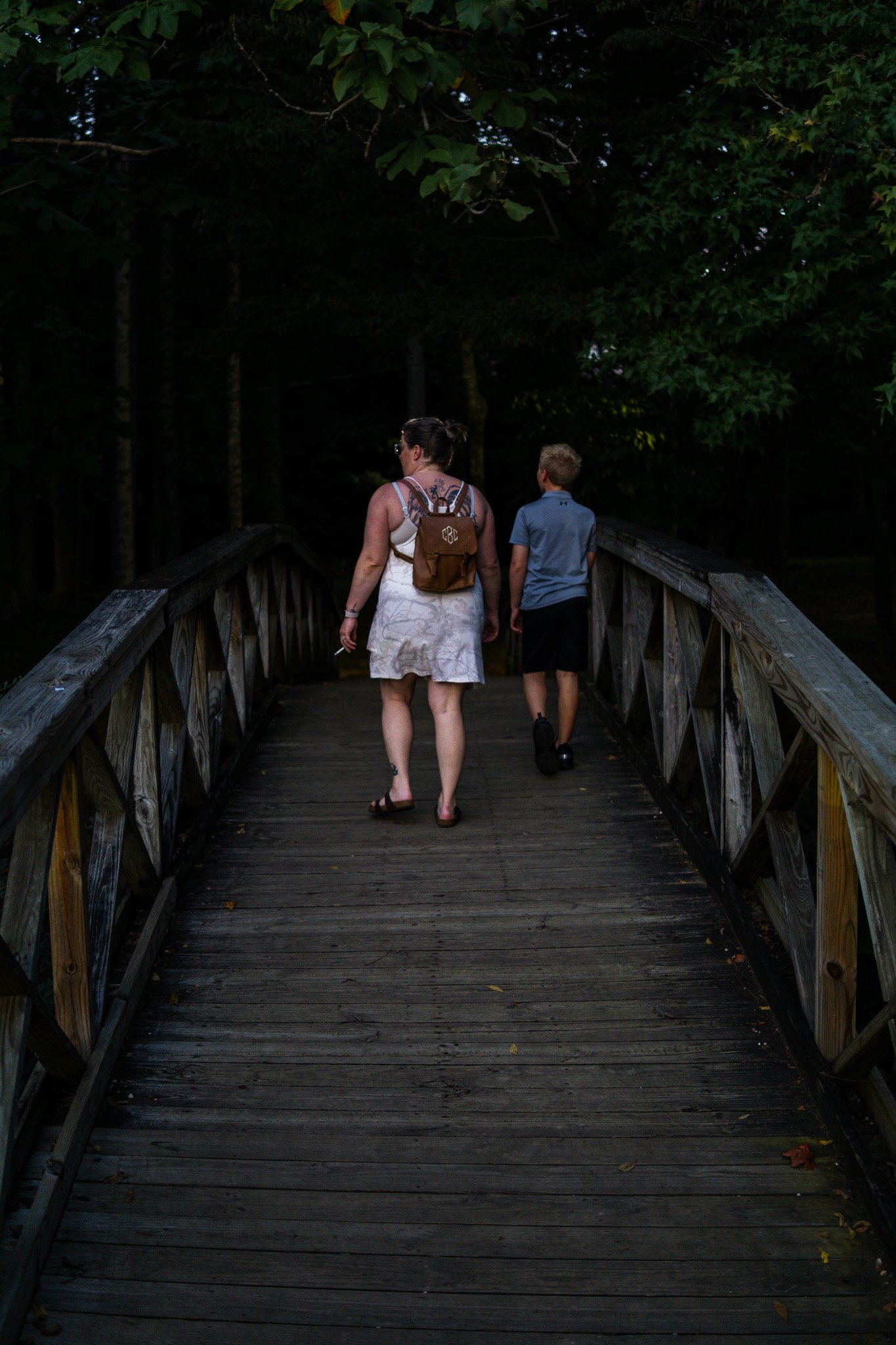 A woman and a young boy walking on a wooden bridge surrounded by trees and greenery.