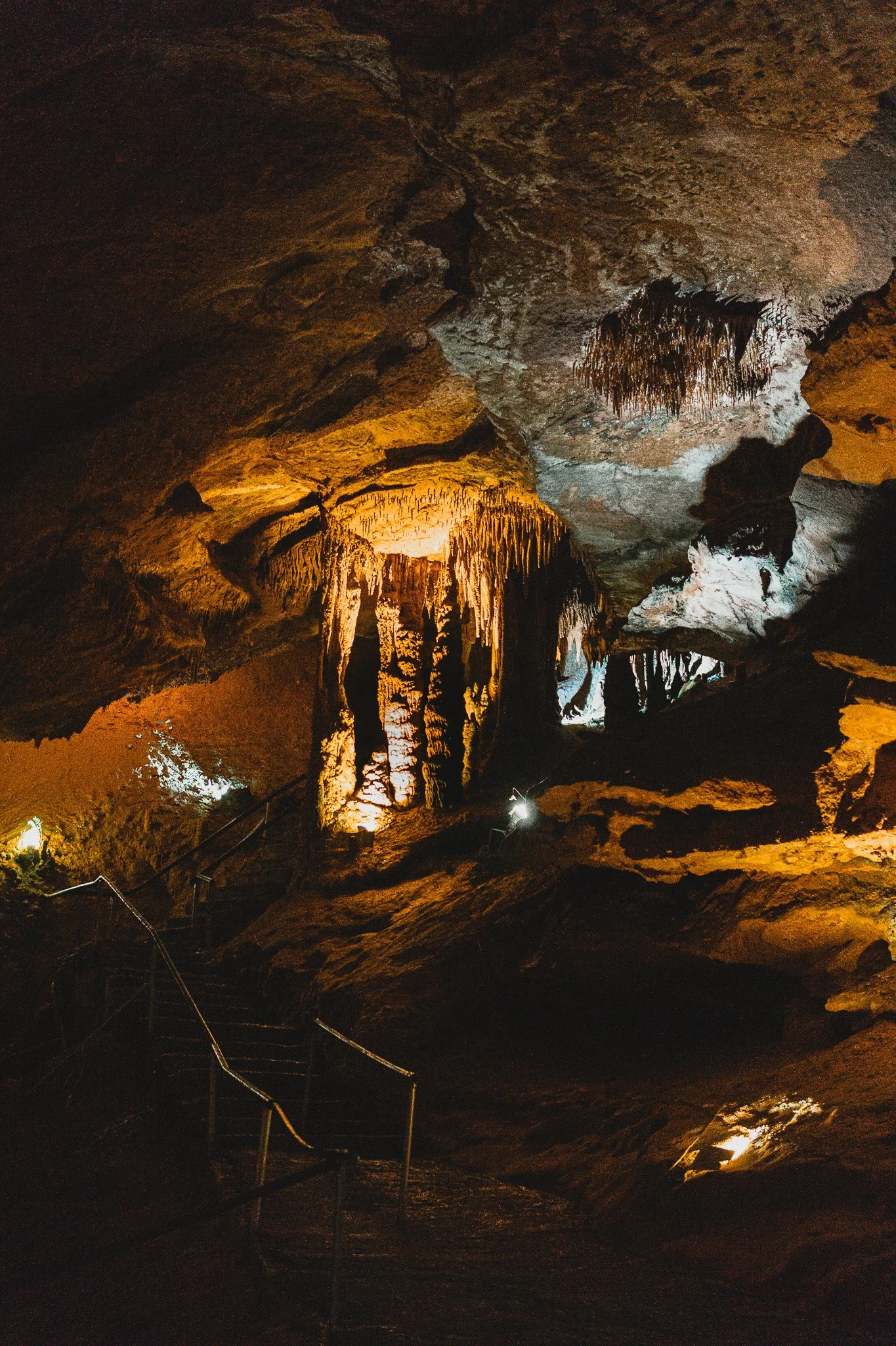 Interior view of a cave with stalactites and stalagmites illuminated by artificial lighting, with a staircase and handrails leading into the cave.