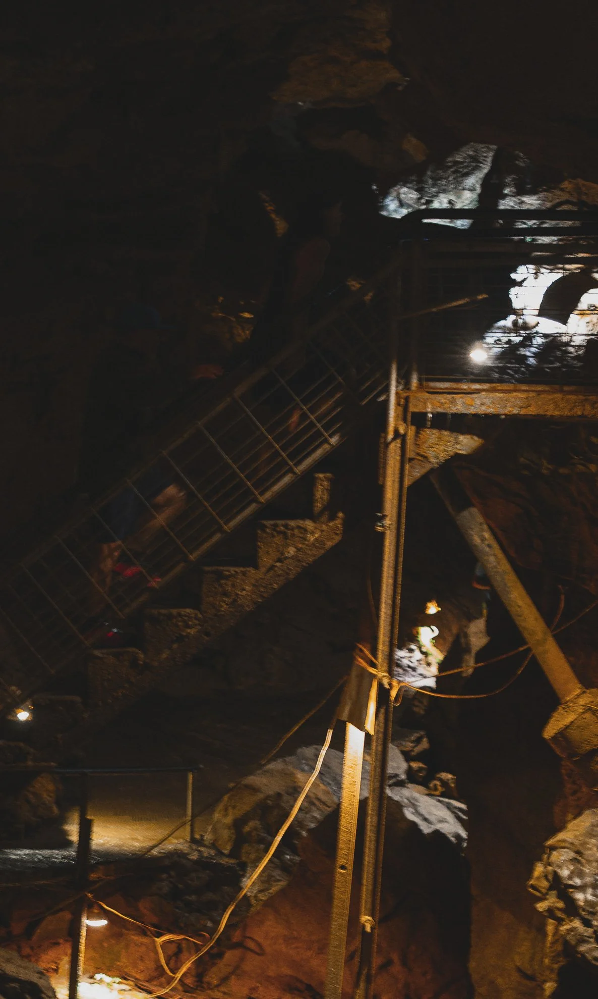 Nighttime scene inside a cave with wooden and metal structures, some rocks illuminated by artificial light, and a person in dark clothing and a helmet climbing the structure.