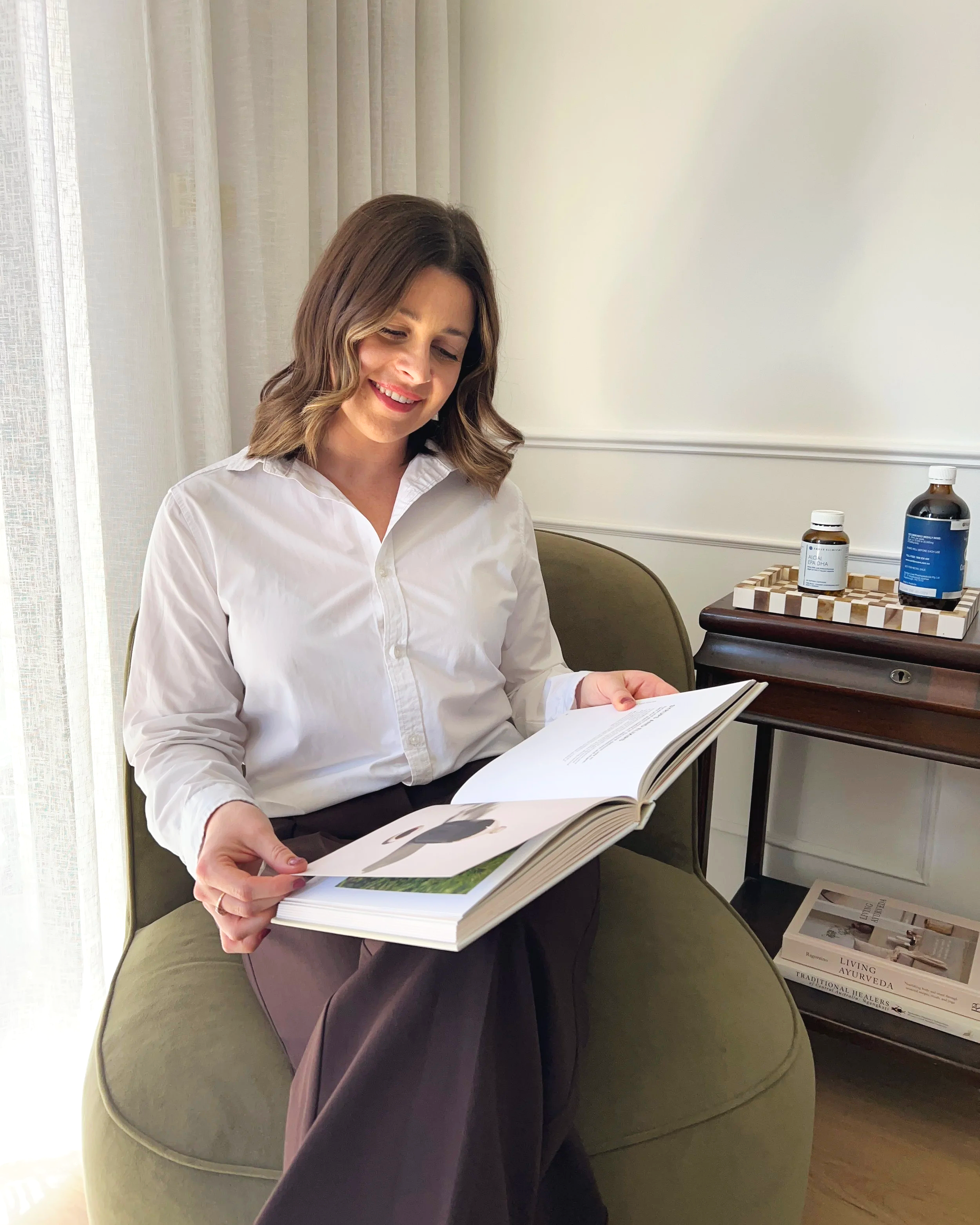 A woman with shoulder-length brown hair and a white button-up shirt sitting on a green curved armchair, reading a large open magazine or book. She is smiling and appears to be in a bright room with white curtains and a table next to her that has bottles and a wooden tray. A stack of books or magazines is on the floor near the table.