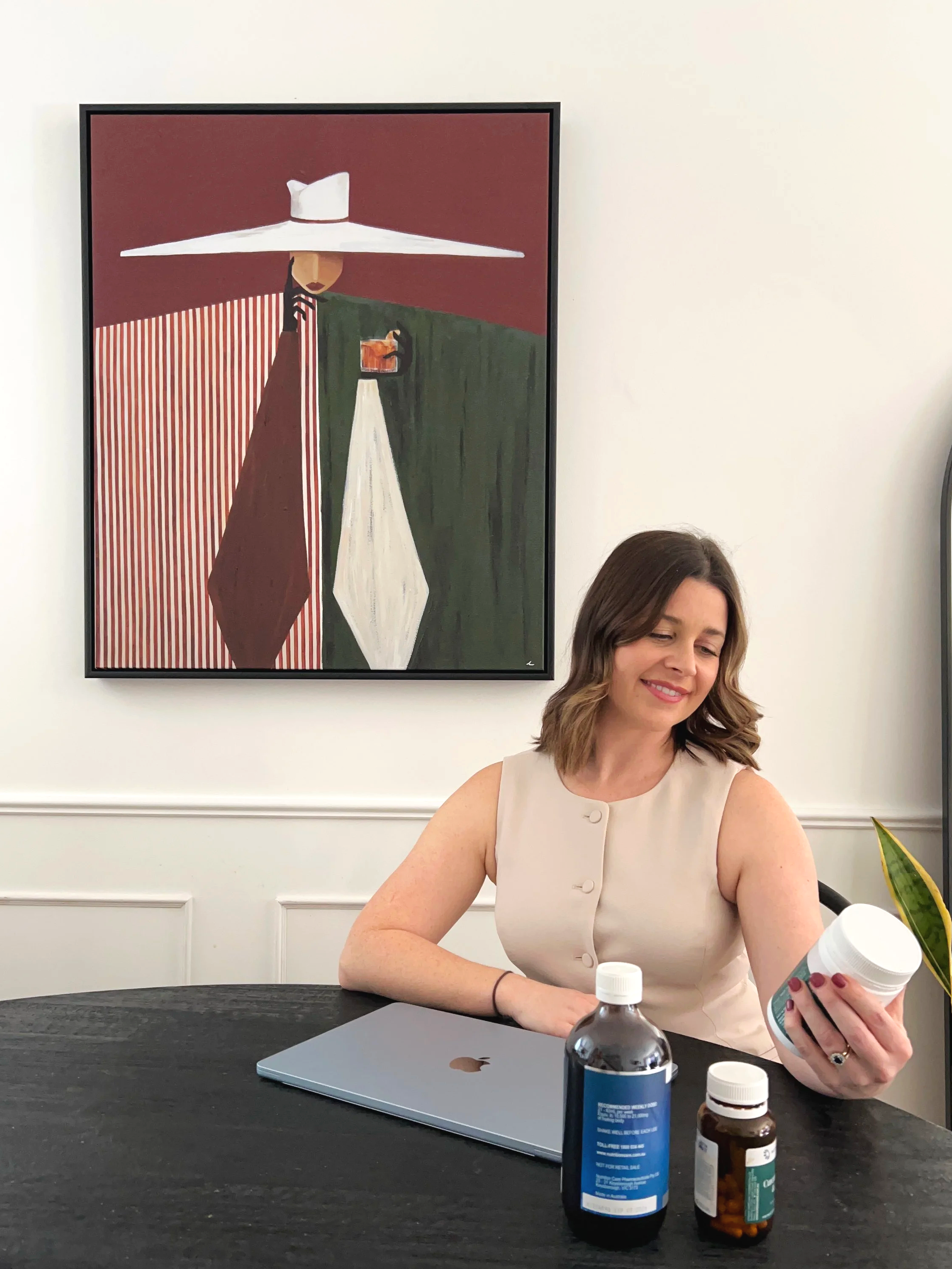 Woman sitting at a table looking at a bottle of pills with a smile. On the table are a water bottle, a laptop, and additional pill bottles. There is an abstract artwork of a woman wearing a large white hat hanging on the white wall behind her.