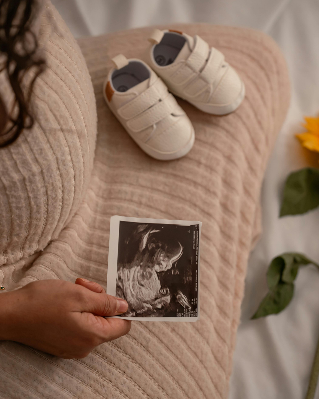 A woman sitting on a beige textured fabric surface, holding an ultrasound image in her hand. On the surface next to her are a pair of small white baby shoes with velcro straps. Part of a yellow flower and green leaves are visible on the right side of the image.