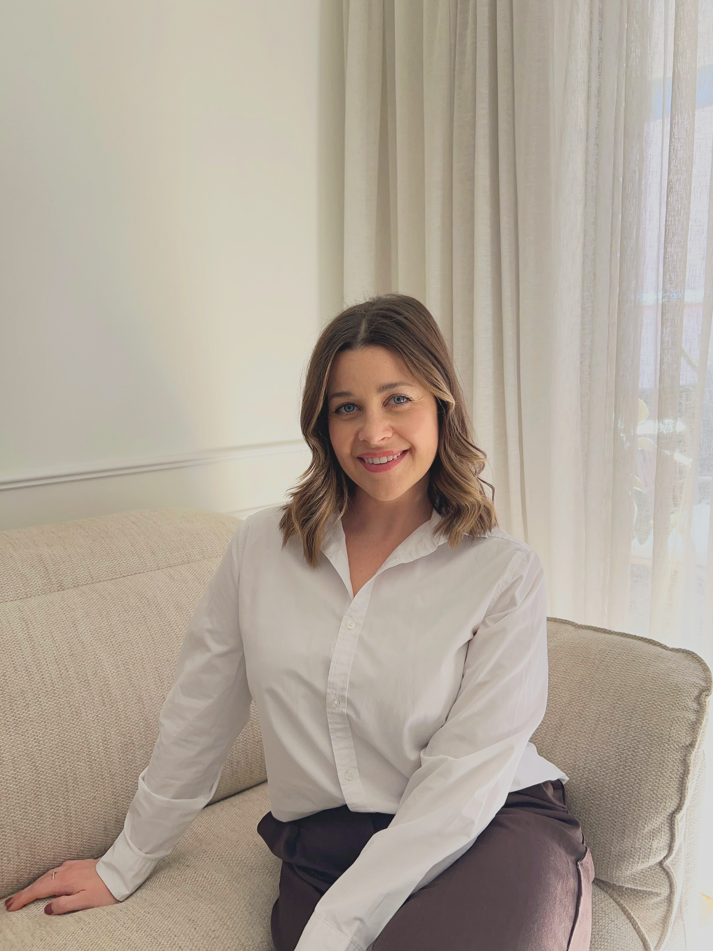A woman with shoulder-length wavy brown hair, blue eyes, and a friendly smile sitting on a beige couch in a well-lit room with cream curtains.