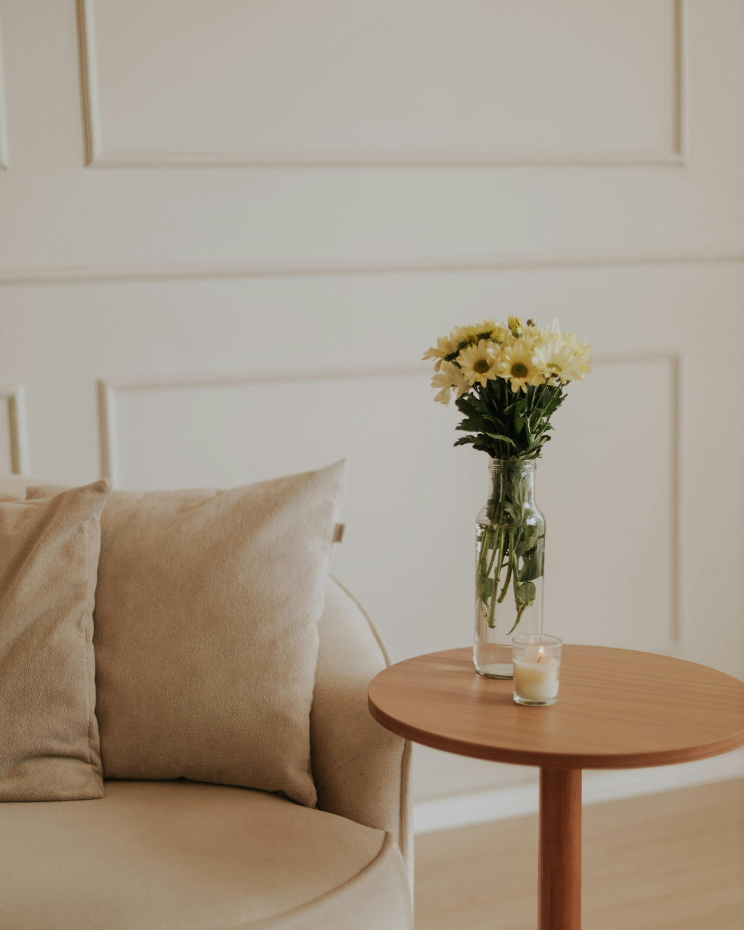 A beige sofa with matching pillows next to a round wooden side table. The table holds a vase with white flowers and a lit candle.