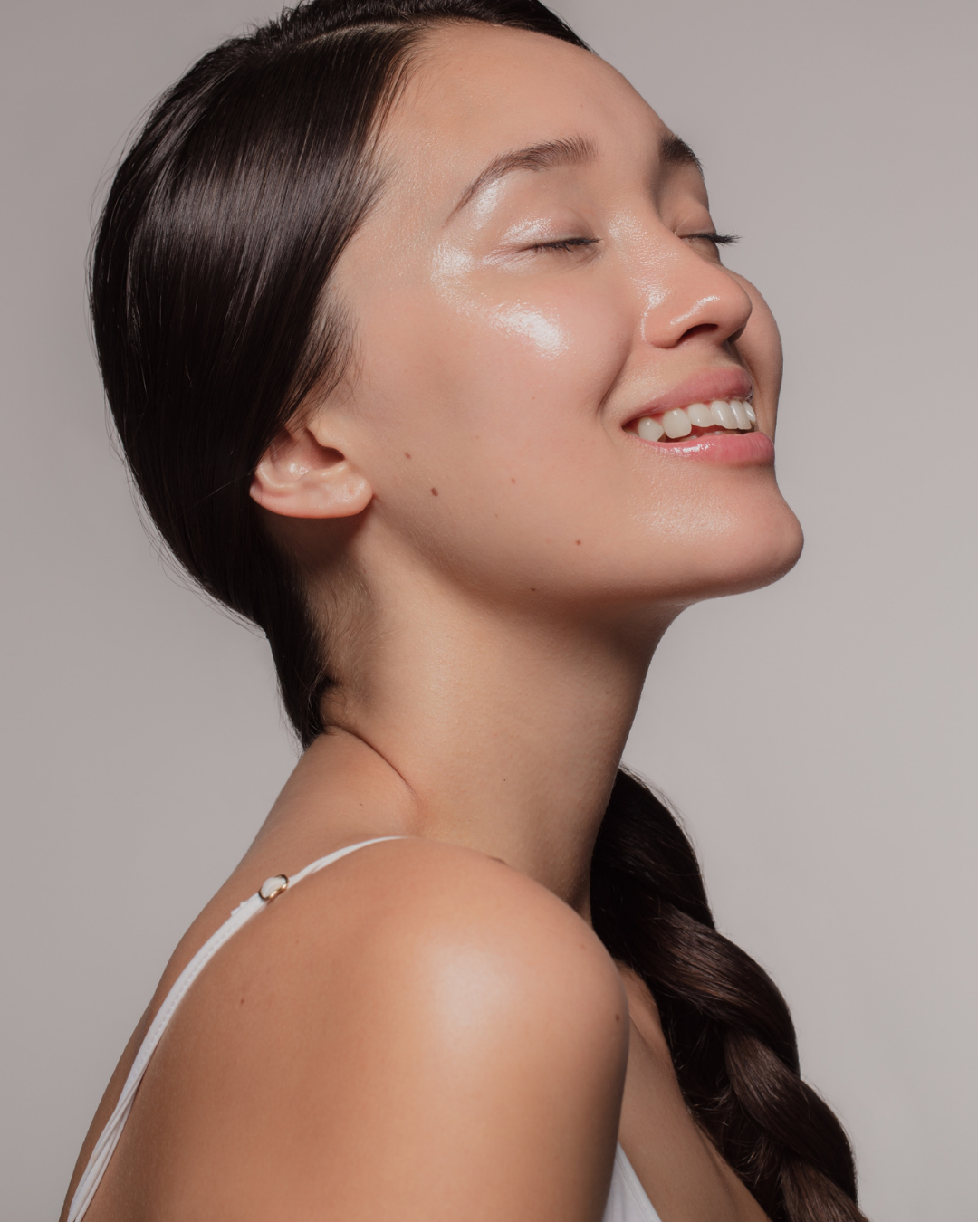 Close-up of a woman with glossy skin, closed eyes, and a smile, with dark brown hair styled in a side braid, wearing a white strap top.