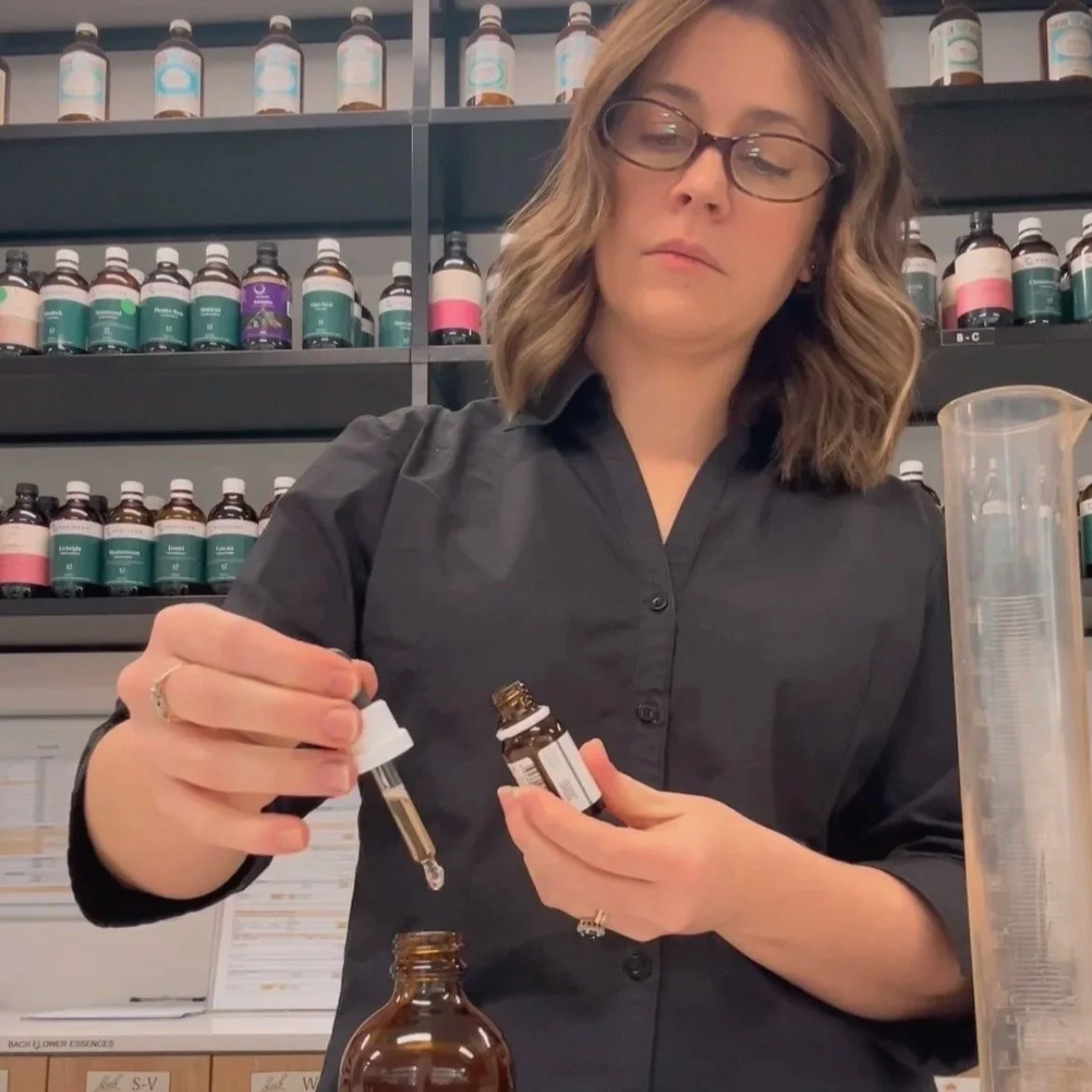 A woman with glasses pouring liquid from a dropper into a small bottle in a pharmacy or apothecary setting, with shelves of various bottles in the background.