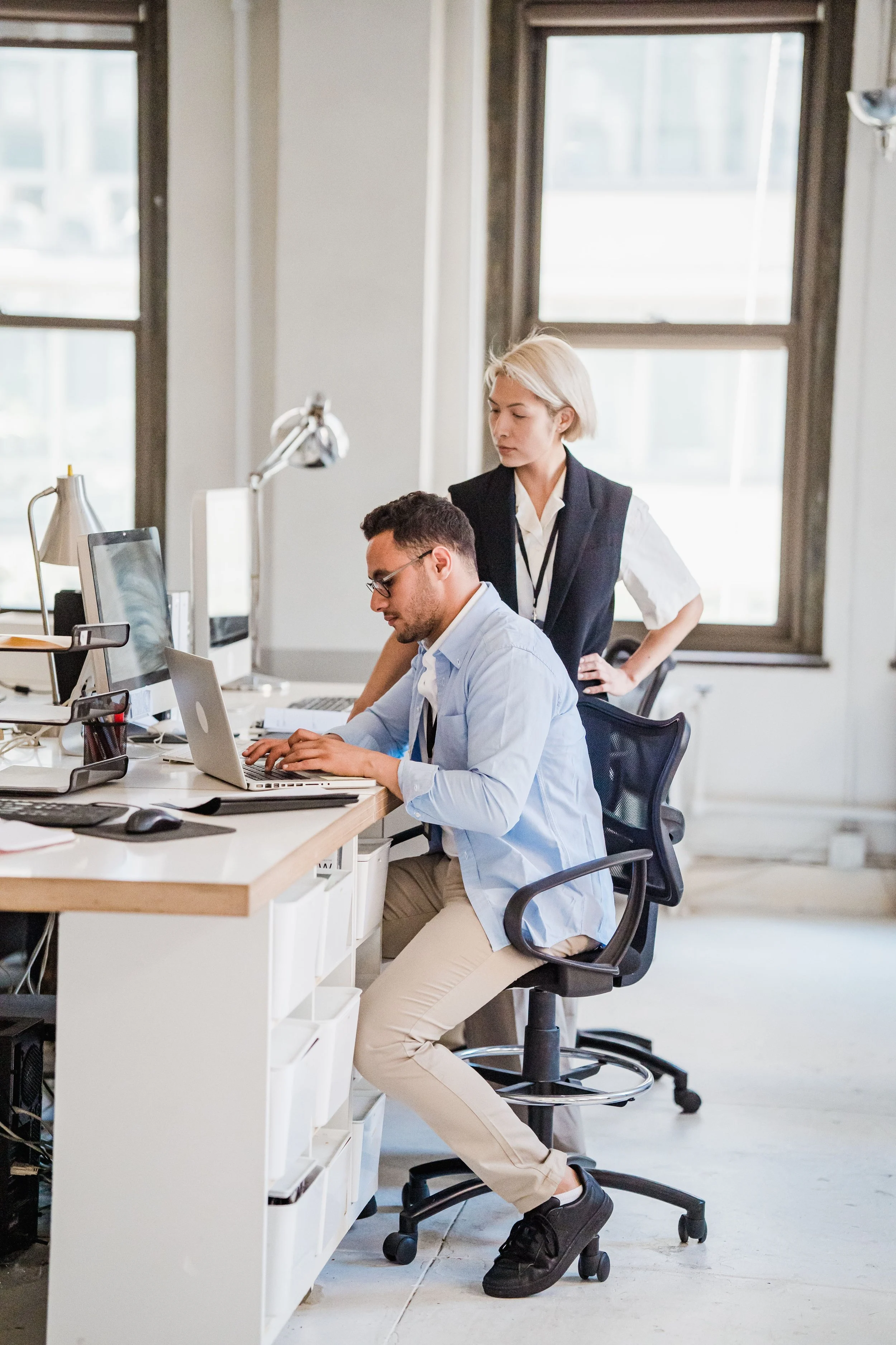 Two coworkers in an office, one woman standing and looking at a computer screen, the other man sitting, working on a laptop at a desk with large windows in the background.