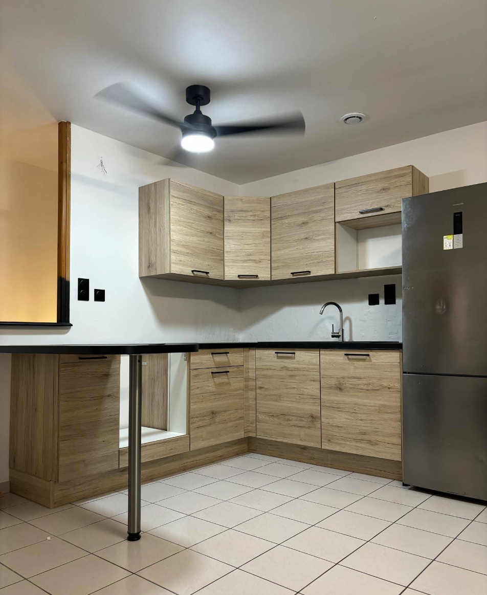 Modern kitchen with light wood cabinets, black countertop, white tiled floor, stainless steel refrigerator, and a ceiling fan.