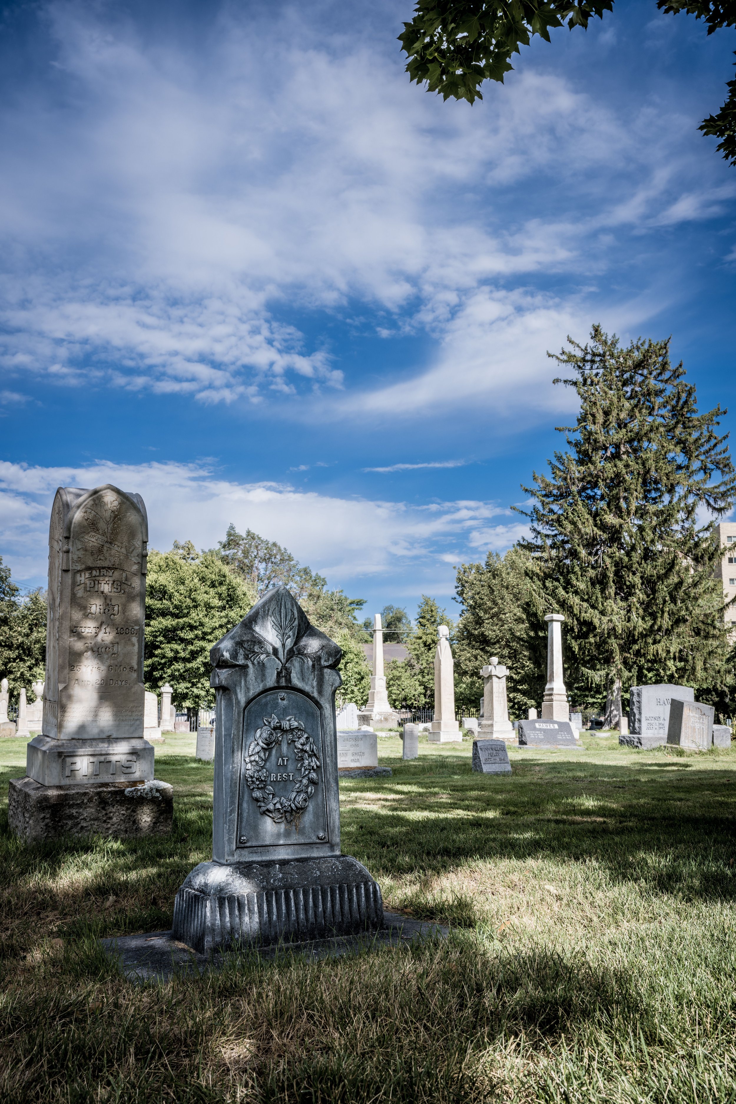 A graveyard with various headstones and monuments, some with flowers and inscriptions, surrounded by green grass and trees under a partly cloudy blue sky.