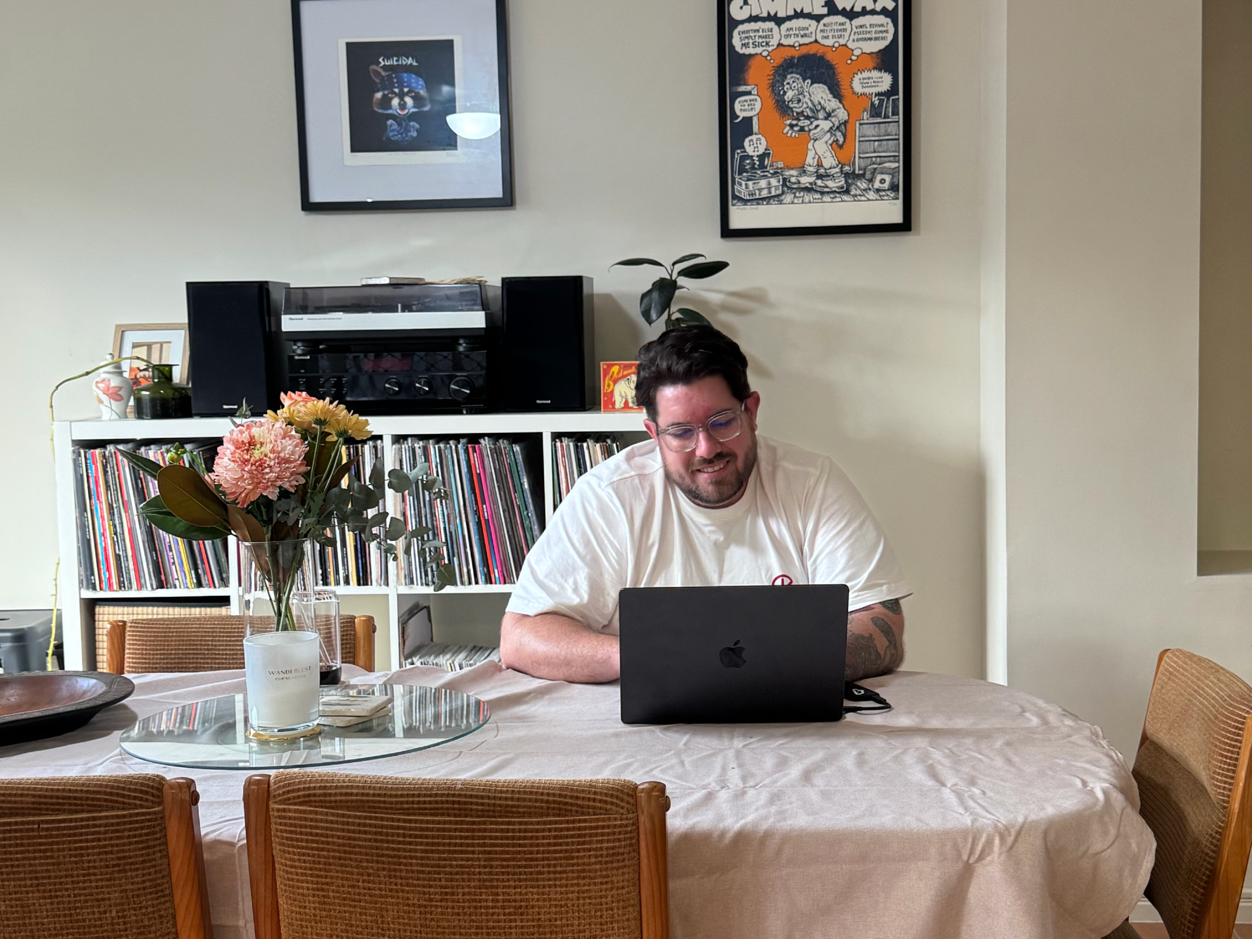 A man with glasses and a beard sitting at a dining table with a laptop in front of him, smiling. The table has a white tablecloth, a glass vase with pink and peach flowers, and a white candle. The background features a bookshelf with records, two framed posters on the wall, and a vintage stereo system.