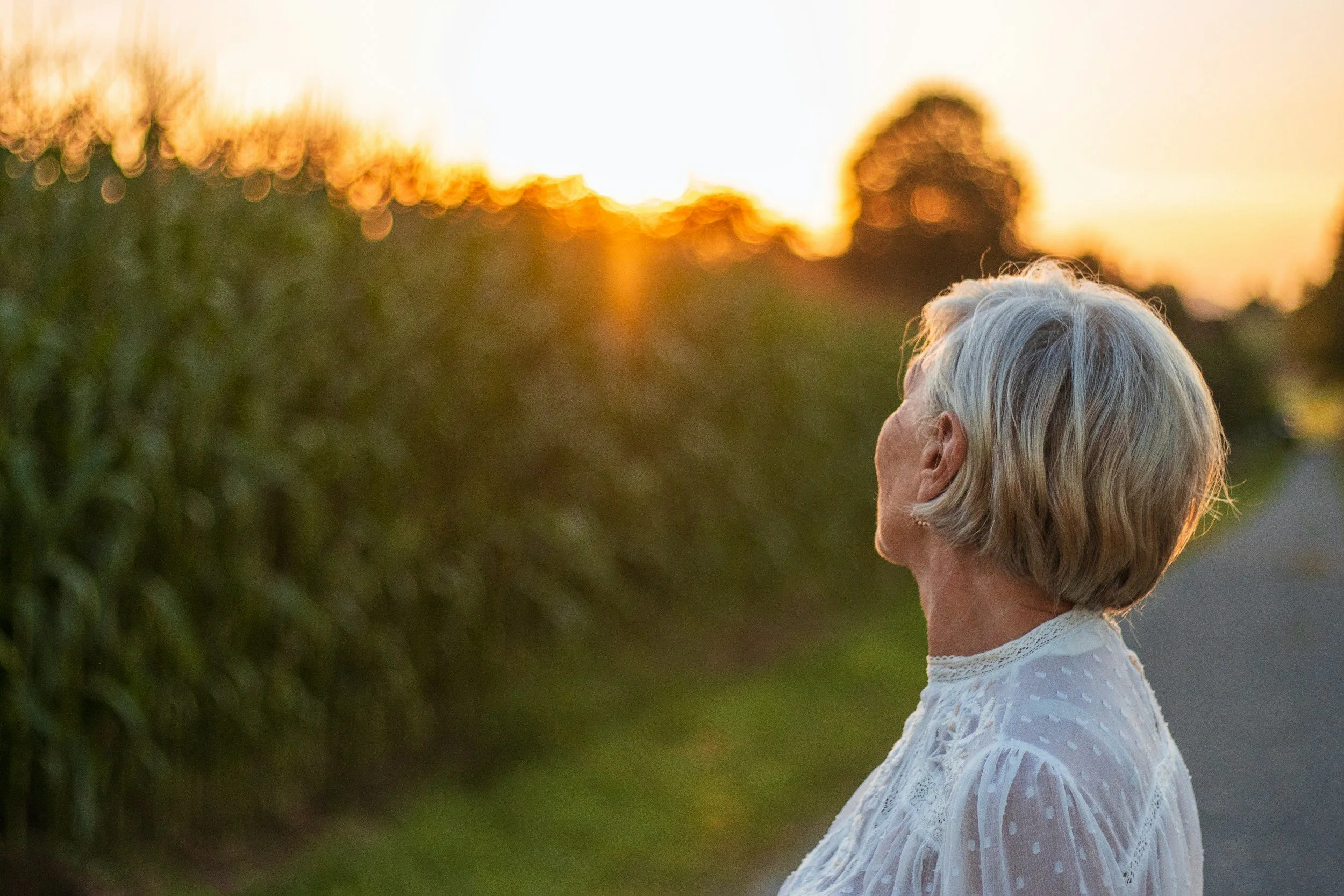 Side view of elderly woman with gray hair wearing a white eyelet dress, standing outdoors during sunset.