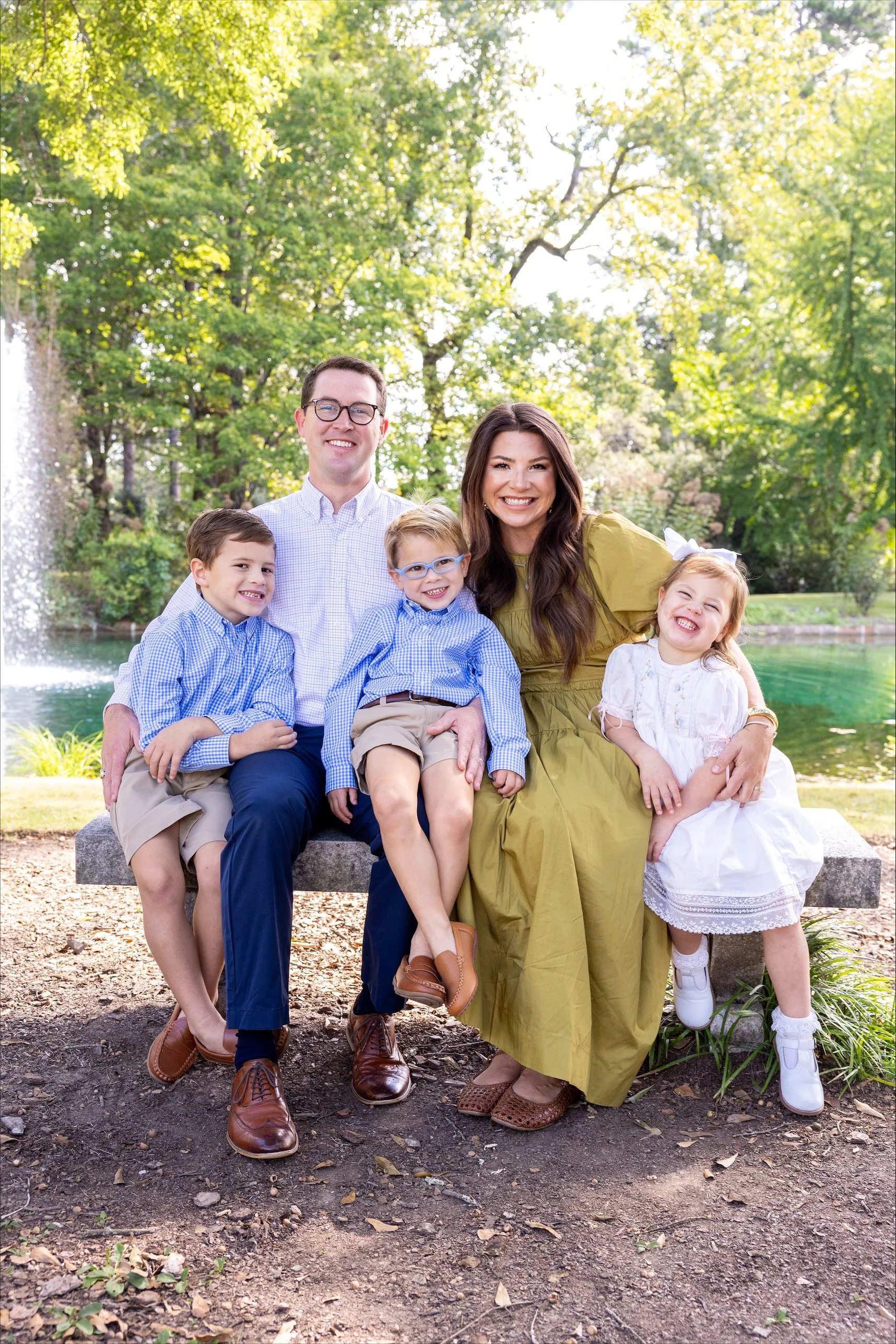 A family of six sitting on a park bench with trees and water in the background, smiling at the camera. The family includes two adults and four children, with the children wearing blue and white clothing.