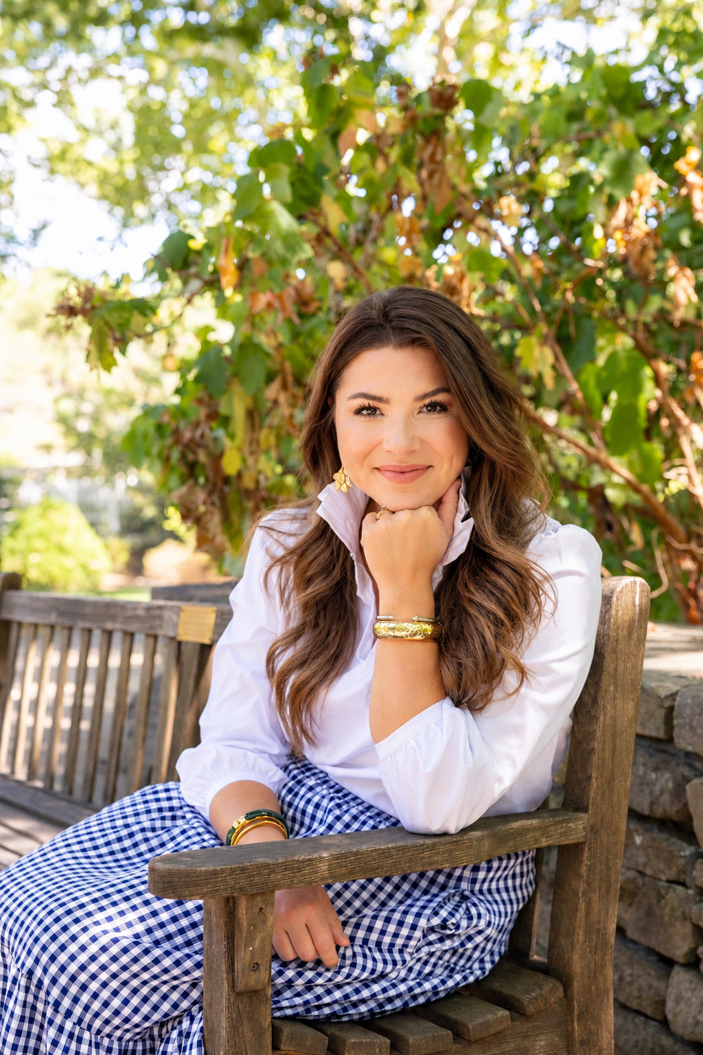 A young woman with long, wavy brown hair sitting on a wooden bench outdoors, smiling gently, wearing a white blouse, gold jewelry, and checkered blue and white skirt, with lush green foliage in the background.