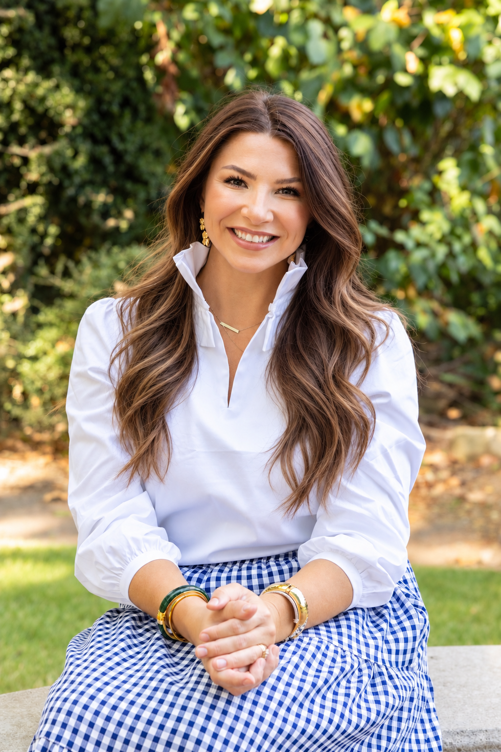 A smiling woman with long wavy brown hair wearing a white blouse and blue gingham skirt sitting outdoors with green trees in the background.