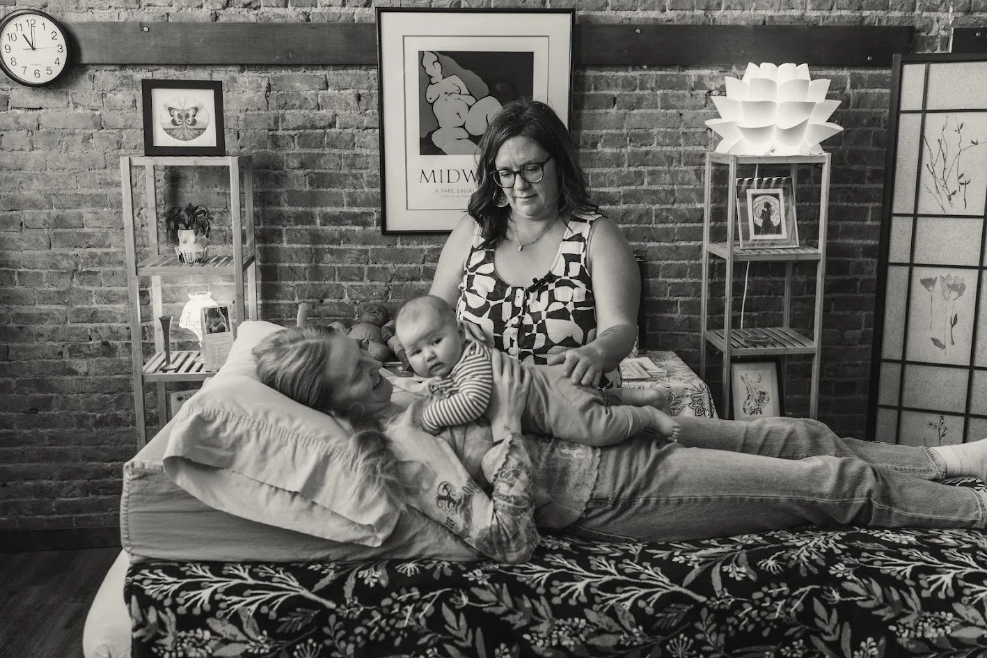 A woman lying on a bed with a pillow, holding a baby on her chest, outdoors with a brick wall background, and another woman standing beside them, touching the baby's leg.