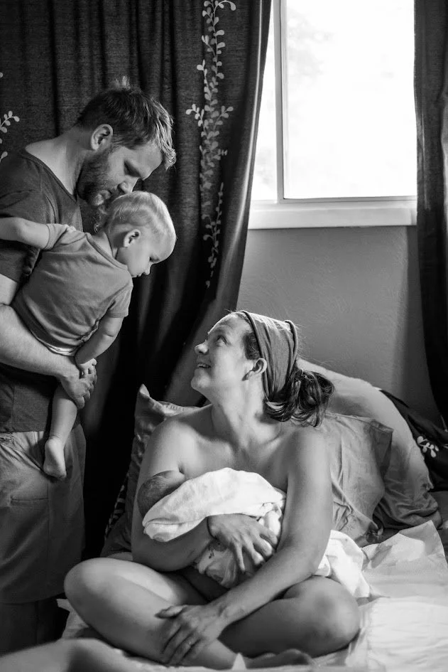 A woman in a hospital bed holding a blanket, smiling at her partner and child who are standing beside her, with a window in the background.