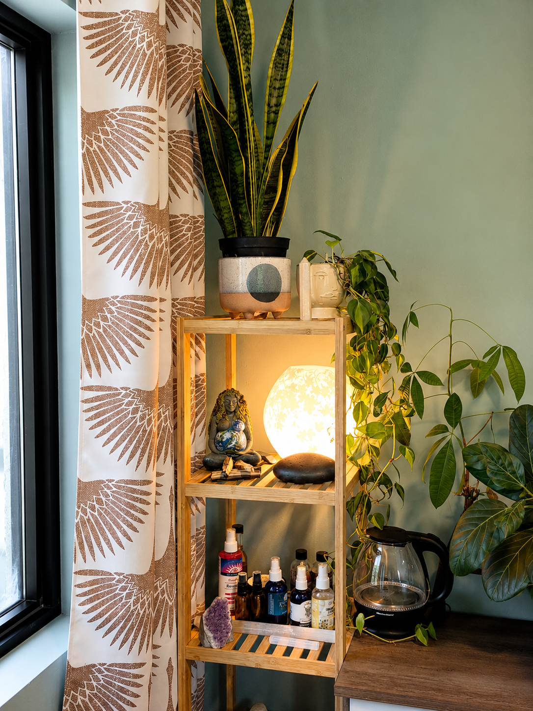 A wooden shelving unit with decorative plants, a lit decorative lamp, a small Buddha statue, and various spray bottles and essential oils, adjacent to a window with patterned curtains and a countertop with a glass coffee maker.