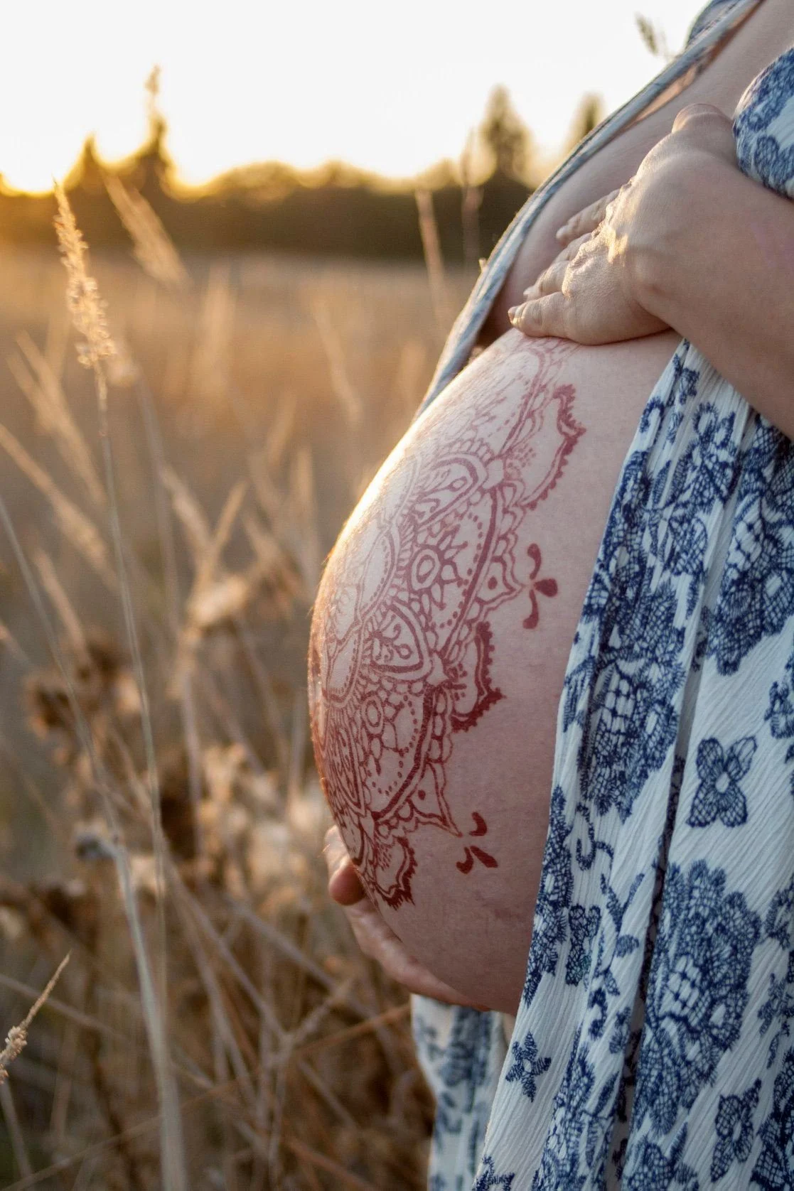 Close-up of a pregnant woman in a field at sunset, with henna designs on her belly and wearing a blue and white printed dress.