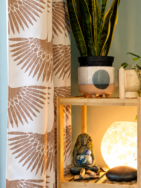 Interior corner with a wooden shelf holding a potted plant, a Buddha figurine, and a glowing table lamp, next to a curtain with a brown leaf pattern.