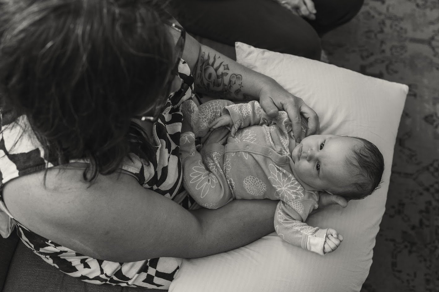 A woman lying on a pillow holds a baby in her arms, looking at the baby who is peacefully resting, wearing a patterned onesie, with a knitted cap, in a cozy setting.