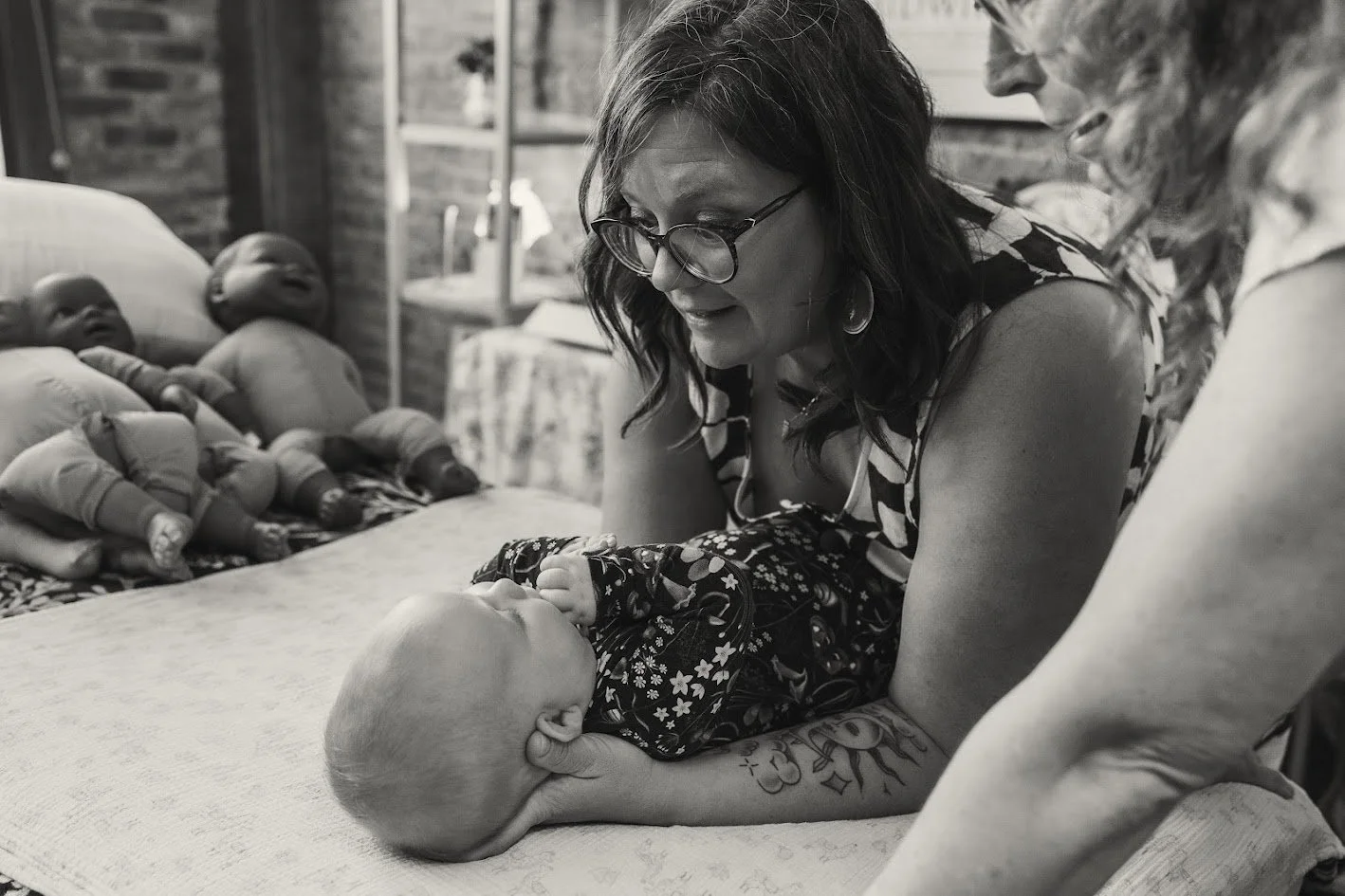 A woman with glasses and tattoos on her arm is looking at a baby lying on a table. The woman is smiling and leaning over the baby. There are several dolls on the bed in the background.