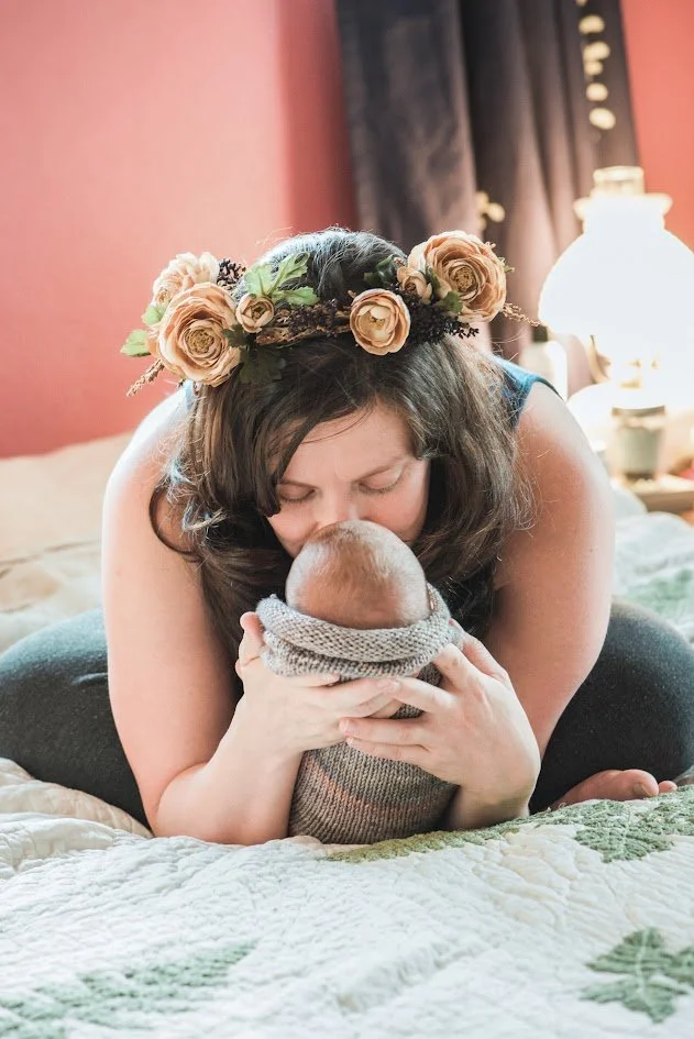 Woman with a flower crown kissing a baby at a bedroom bedside.