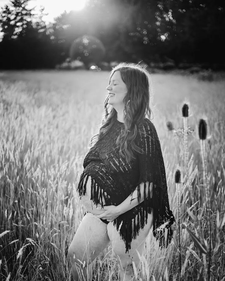 A woman with long wavy hair smiling with her eyes closed, standing in a wheat field during sunset, wearing a black fringed shawl.