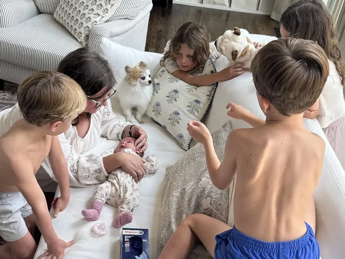 A group of children and a woman are gathered around a newborn baby on a couch. Some children are petting the baby and dogs, and one child is shirtless and sitting on the floor. A box of baby formula is visible on the couch near the woman.