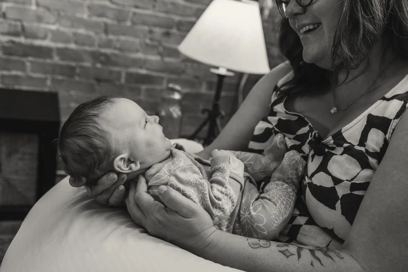 A woman holding a young child on a bed, looking lovingly at the child in a cozy, indoor setting with a brick wall and a lamp in the background.